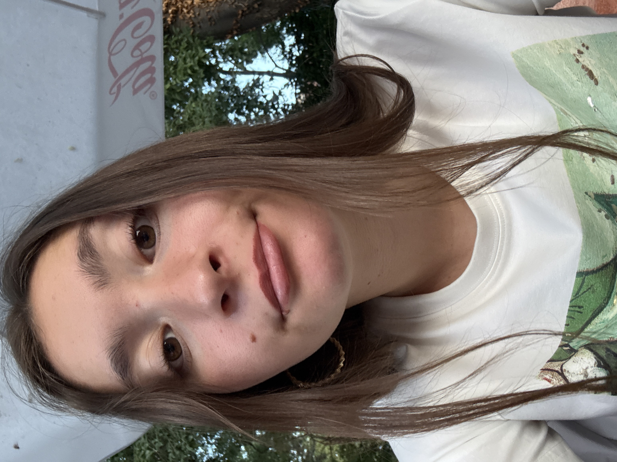 This is a close-up, rotated selfie of a young woman with long, dark brown hair and brown eyes. She is wearing a white t-shirt with a green and brown graphic print. Her lips are slightly parted, and she is looking directly at the camera with a neutral expression. A small mole is visible on her left cheek, and she is wearing a gold hoop earring.
In the background, a portion of a white object with a pink logo is visible on the left, which appears to be a Coca-Cola logo. Beyond that, green foliage and trees are visible, suggesting an outdoor setting. The lighting indicates it could be daytime. The image appears to have been taken in Sozopol, Bulgaria.