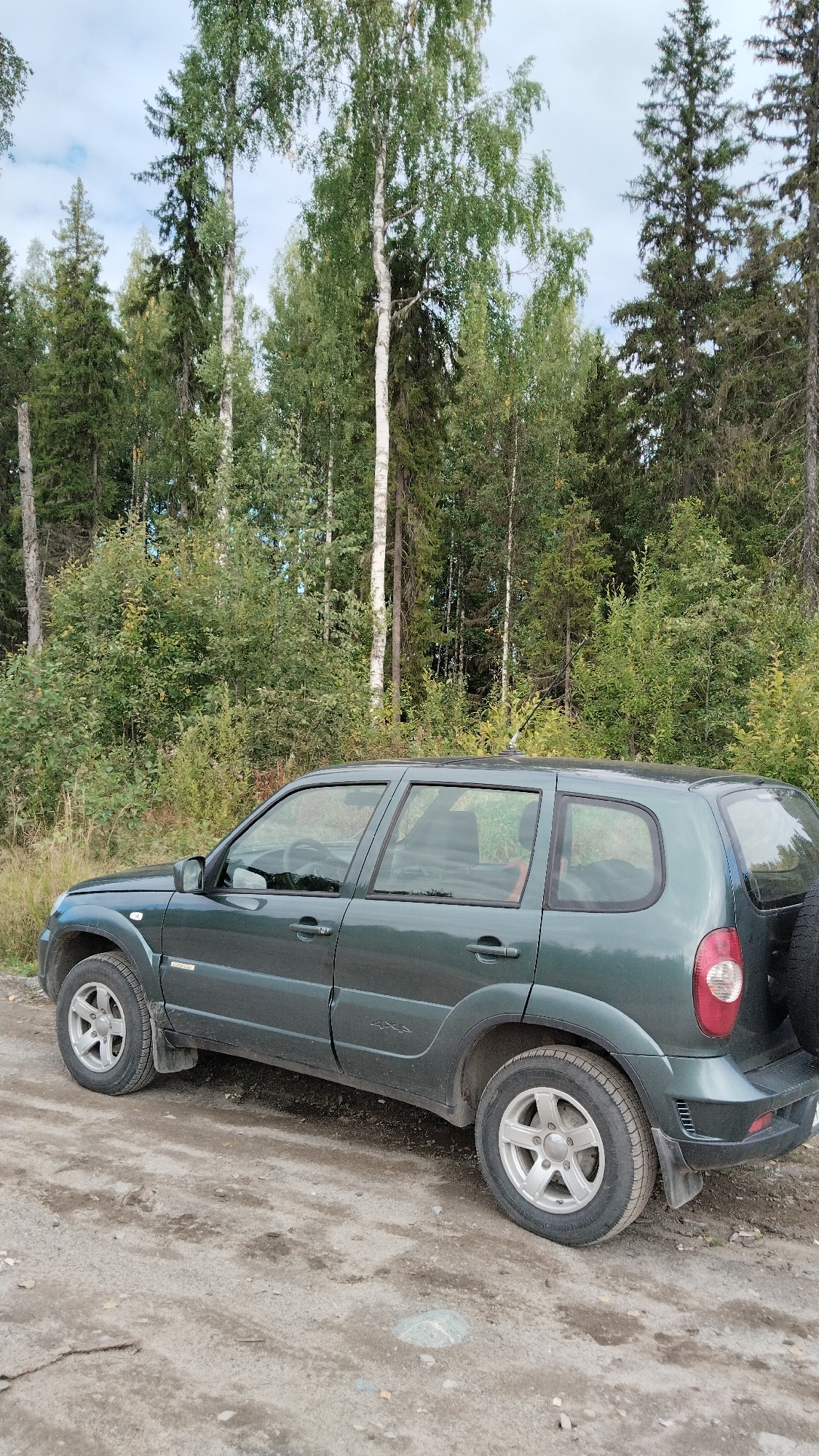 A dark green, four-door SUV is parked on a dirt road. The vehicle is viewed from the side, with the front of the car on the left. The SUV has alloy wheels and a spare tire mounted on the back. Behind the car is a dense forest with a mix of deciduous and coniferous trees. The sky is overcast with some patches of blue visible. The overall scene suggests a remote, natural setting.