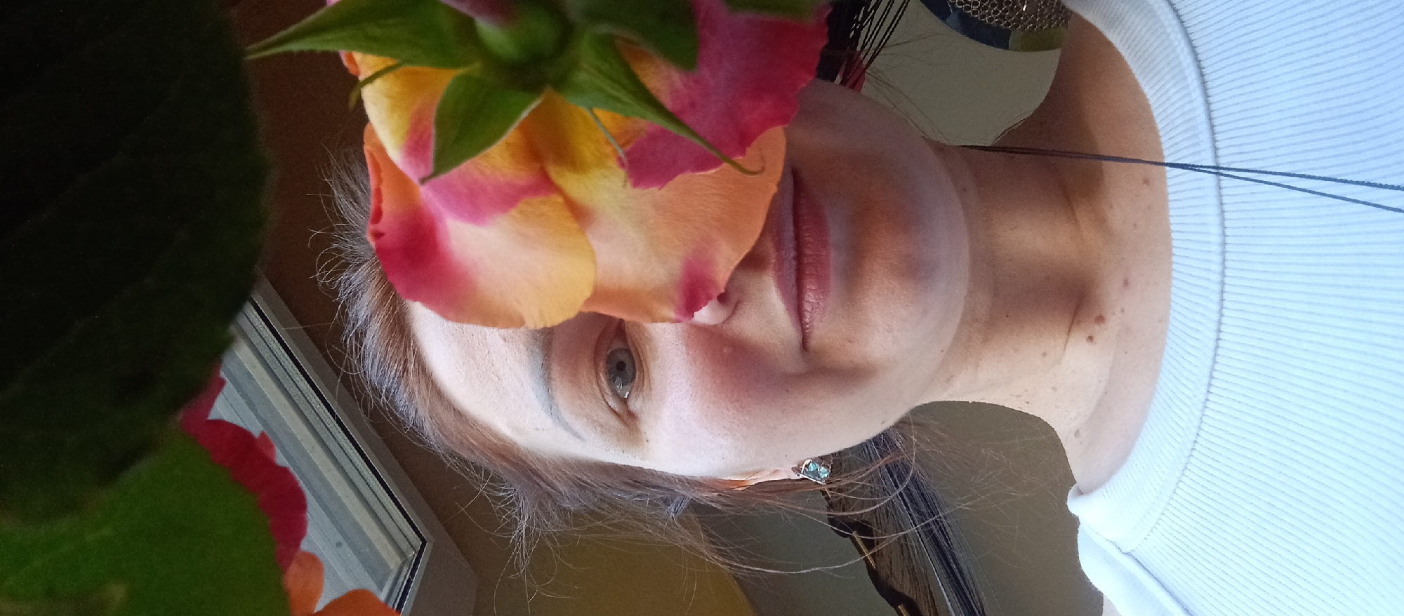 A close-up, angled shot depicts a person's face partially obscured by a bicolor rose. The person has fair skin, light-colored eyes, and light brown hair. They wear a light-colored, ribbed top. The rose, positioned near the top of the frame, is primarily yellow and peach with pink tips. A green, leafy plant appears in the lower left corner. A window and other objects are visible in the background. The location is Unknown, Unknown.