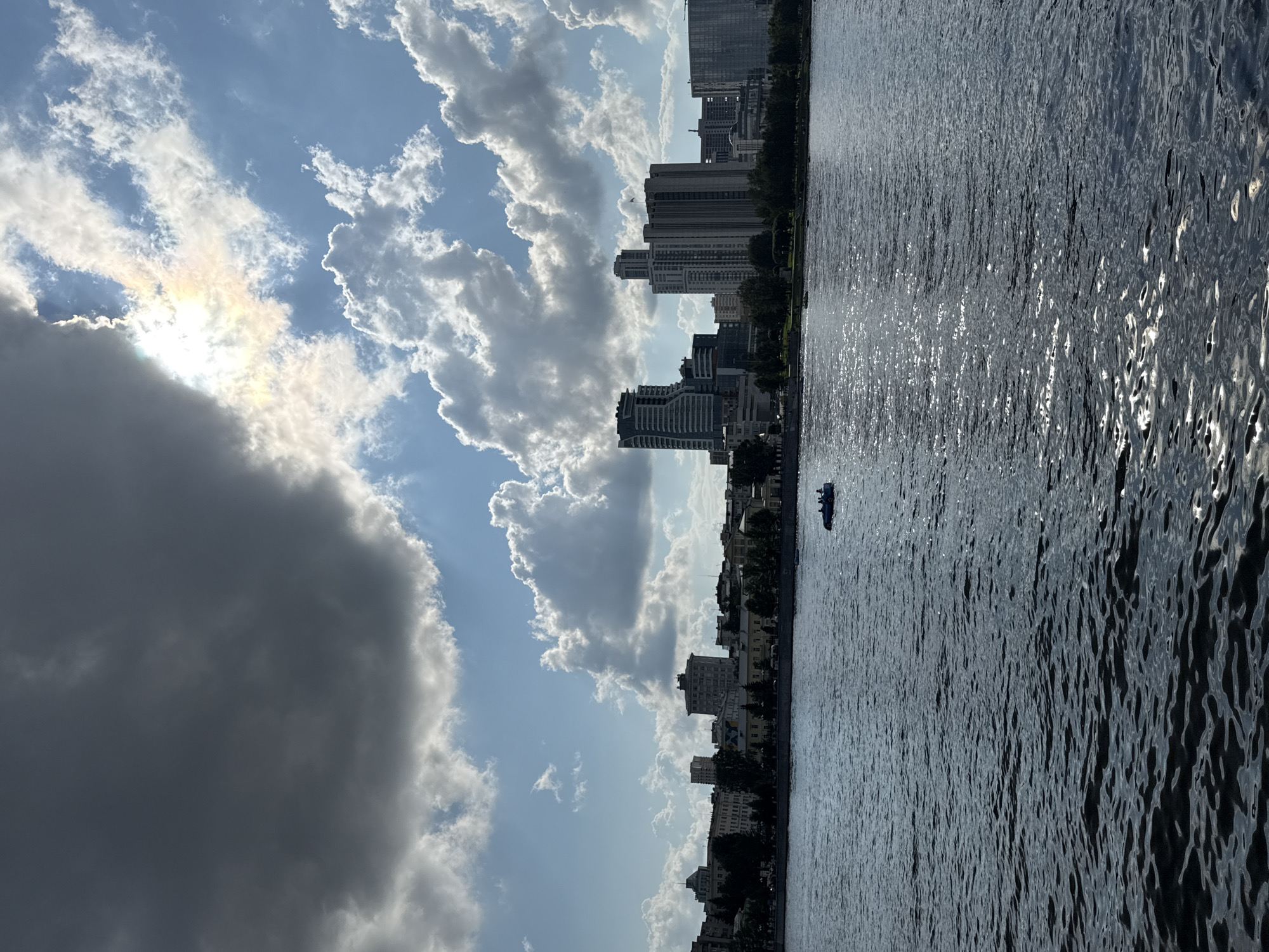 The image shows a wide shot of the city of Yekaterinburg, Russia, on a cloudy but bright day, with a river in the foreground. The river water is dark and reflects the sunlight in shimmering patches. In the middle distance, a small boat with several people on board is visible on the water. The city skyline is dominated by a row of buildings along the riverbank, including a cluster of modern high-rise buildings with glass facades. Trees line the riverbank in front of the buildings. The sky is a mix of blue and dramatic dark clouds, with the sun peeking through and creating a rainbow effect in the clouds.