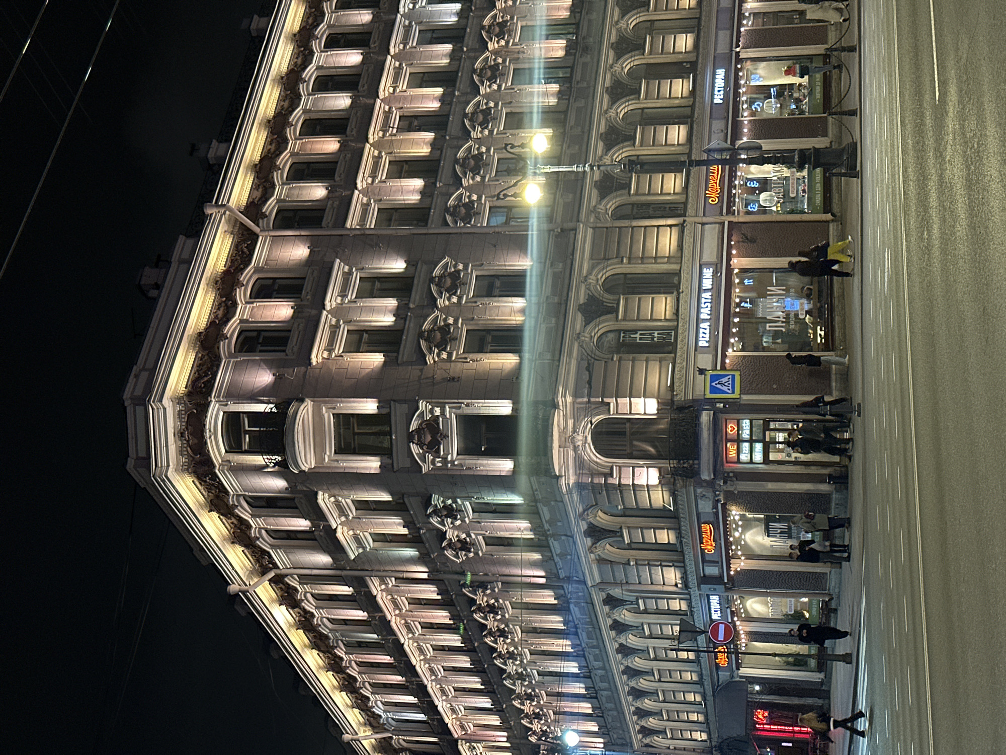 The image depicts a building facade and a street scene at night, likely in Saint Petersburg, Russia. The architecture of the building is ornate, with many windows and decorative elements, illuminated by warm lighting. Streetlights are visible and cast reflections on the wet pavement, indicating possible recent rain.
Several pedestrians are present on the sidewalk, some standing in front of the building's storefronts. The storefronts feature signage, including "РЕСТОРАН," "Марчелли," and "PIZZA PASTA WINE" in Russian and English, suggesting restaurant and dining establishments. A pedestrian crossing sign is also visible, indicating pedestrian infrastructure.