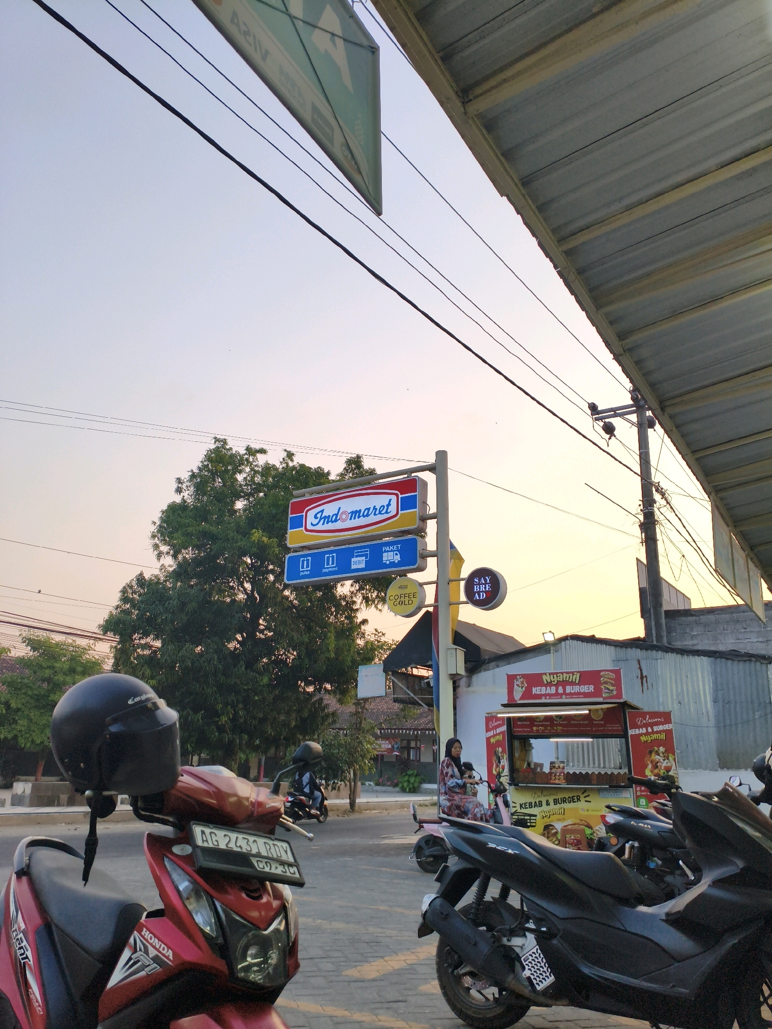 The image depicts a street scene at dusk in Tegalrejo, Indonesia. In the foreground, a red Honda motorcycle with a black helmet on its handlebars is parked. To the right, a black scooter is also parked. Behind the motorcycles, a food stall branded "Nyamil Kebab & Burger" is visible, with a woman sitting on a pink scooter in front of it. Above the stall, an "Indomaret" convenience store sign is prominent, along with other signs including "Say Bread" and "Coffee Gold." The sky is illuminated by the setting sun, casting a warm glow over the scene. Power lines crisscross overhead, and a large tree is situated behind the Indomaret sign. The ground is paved, and buildings line the street in the background.