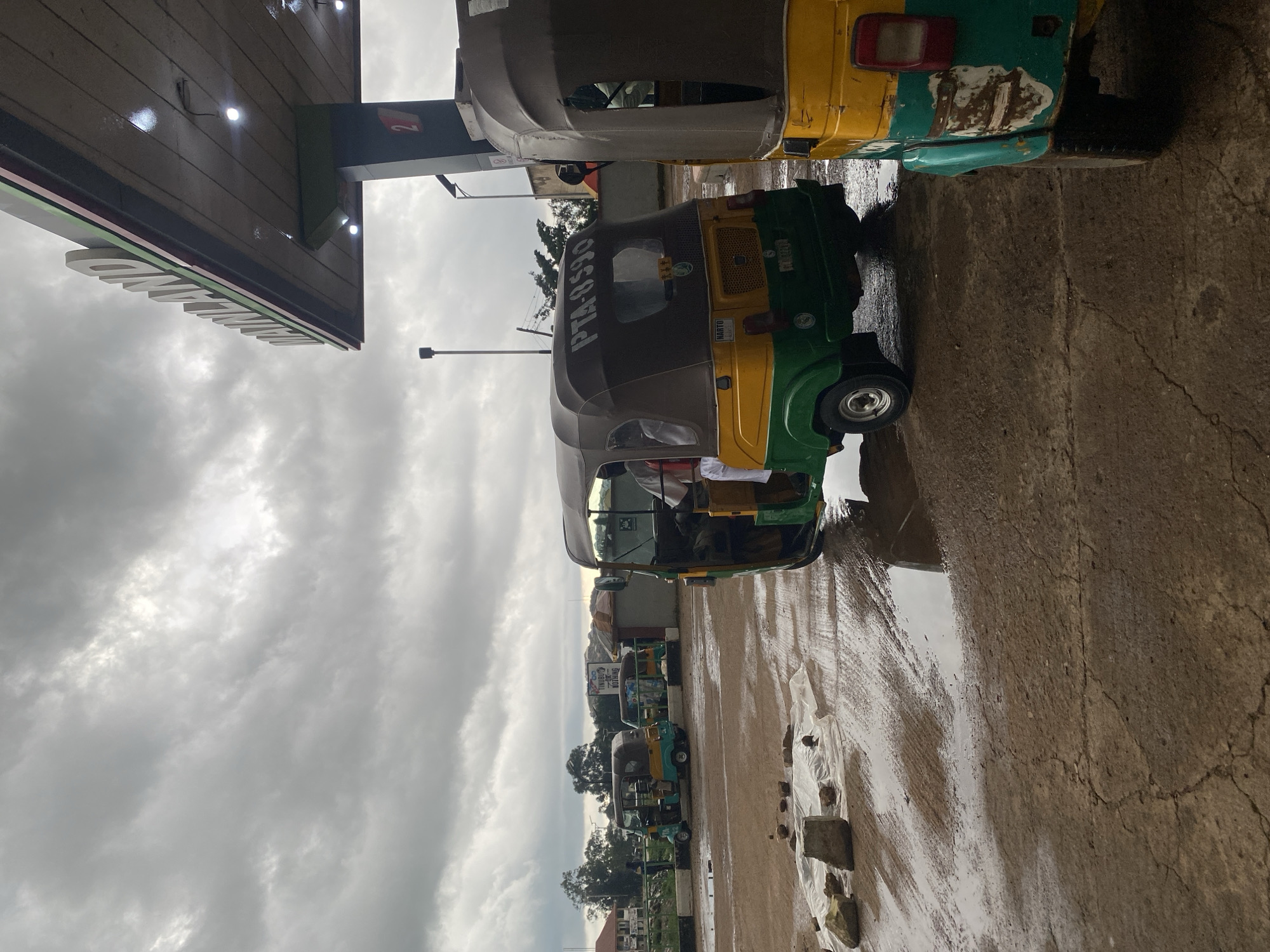 In Gura Top, Nigeria, a gas station with a "MAINLAND" sign is visible under a cloudy sky. A yellow and green auto-rickshaw, license plate PTA-8598, is parked under the station's awning. Another similar vehicle is partially visible on the right. The ground is wet, with puddles reflecting the overcast sky. Rocks and a white plastic sheet are scattered on the wet ground in the foreground. In the background, more auto-rickshaws are parked and trees and buildings are visible.