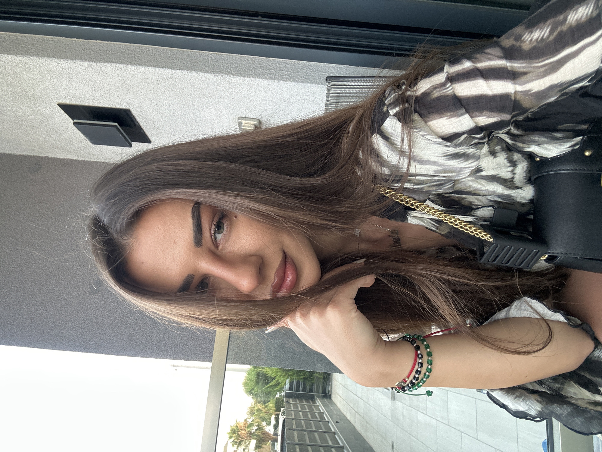 In a selfie taken on a balcony overlooking a residential area with palm trees and manicured hedges, a young woman with long, dark brown hair and makeup is posed. She is leaning on her hand, with her left arm adorned with bracelets. She wears a patterned blouse and a black purse with a gold chain strap rests on her lap. The background features a textured gray wall and a glass door.