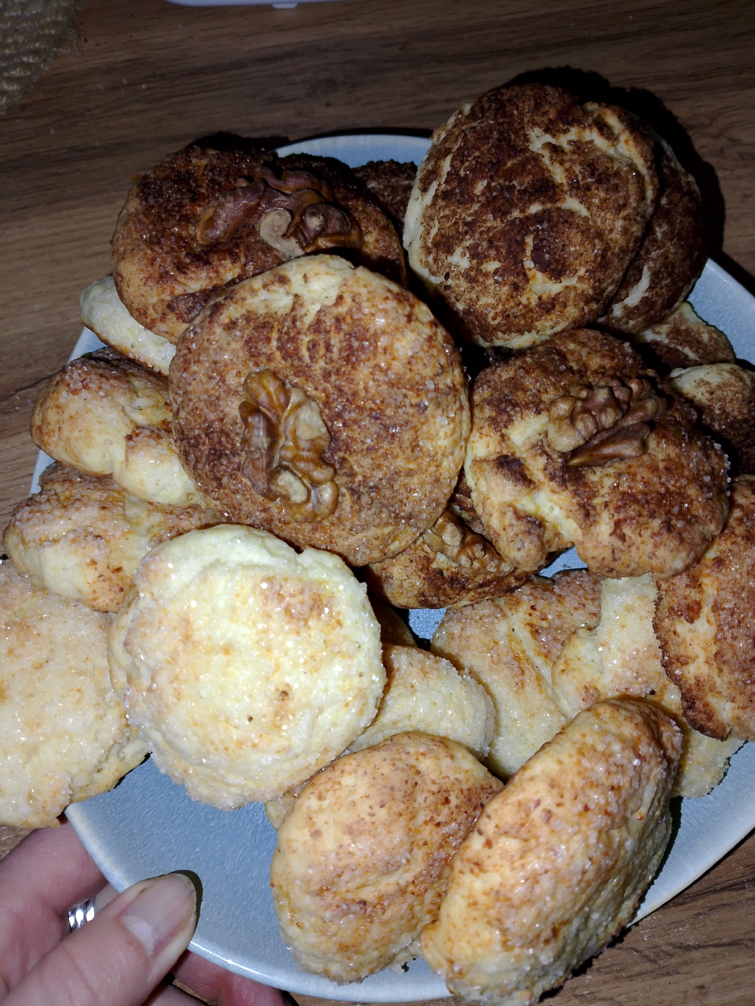 A blue plate piled high with assorted cookies sits on a wooden surface. Some cookies are round, covered in sugar, and topped with a walnut half. Others are coated in a brown speckled pattern. A hand with a wedding band holds the edge of the plate. The location is not specified in the image and there is no evidence that the scene is in Izhevsk, Russia.