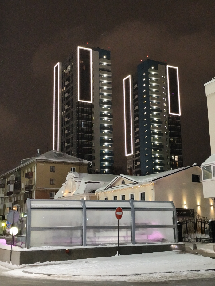 Two tall modern buildings dominate the nighttime scene, each featuring a light rectangle near the top. The buildings have numerous illuminated windows. Below, several older buildings with snow-covered roofs are visible. A long, low structure with a translucent wall runs across the front, partially obscuring the older buildings. A round street lamp and a "Do Not Enter" sign stand in front of the structure, all situated on a snow-covered ground.