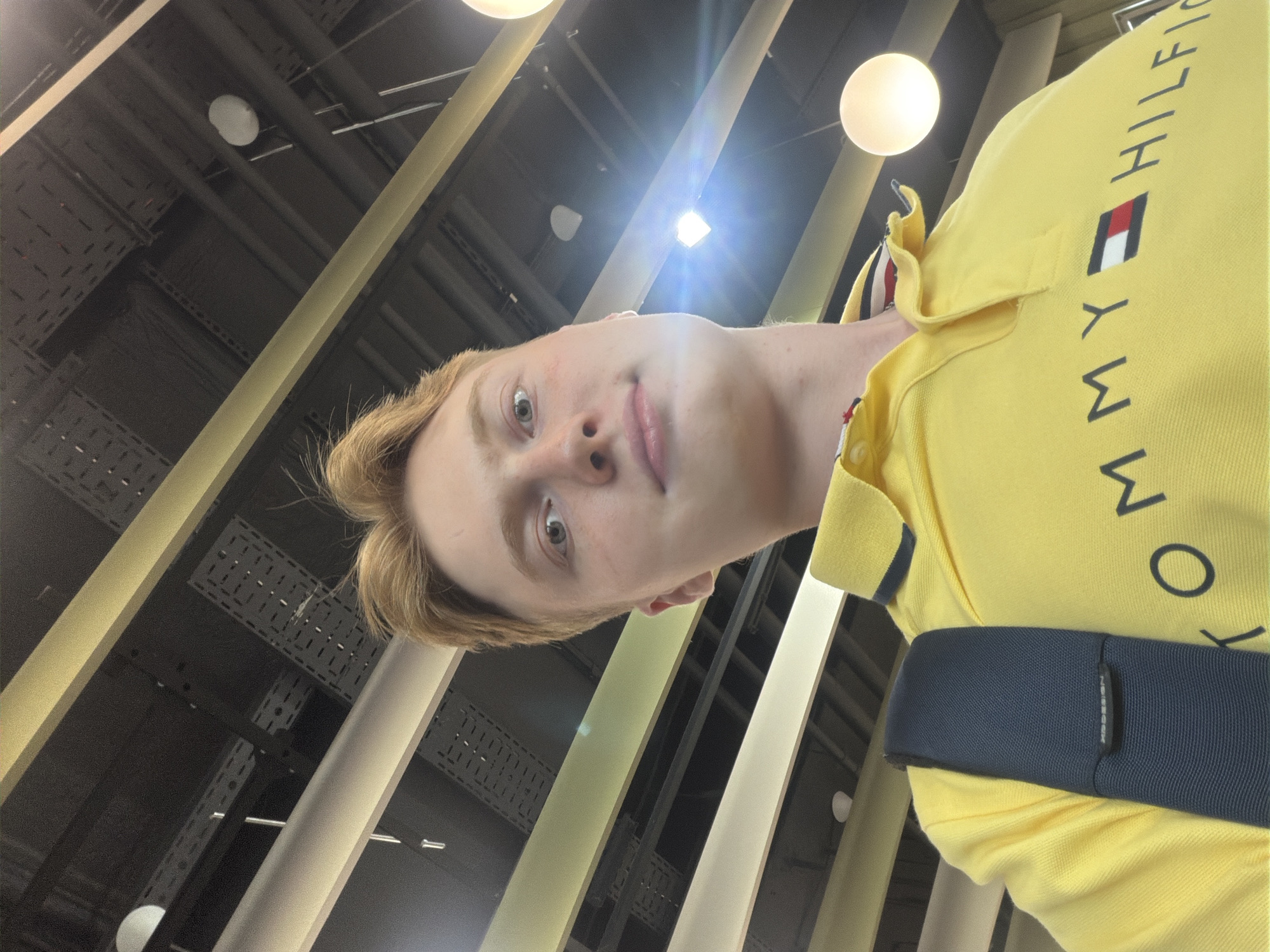 A young Caucasian male with blonde hair takes a selfie in a dimly lit indoor space with a ceiling featuring exposed industrial elements and yellow and white beams. He is wearing a bright yellow polo shirt with a navy collar and the "TOMMY HILFIGER" logo across the chest. A navy backpack strap is visible across his left shoulder. The background lighting includes bright white circular fixtures. There is no information in the image to suggest the location is the city of Kyiv, Ukraine.
