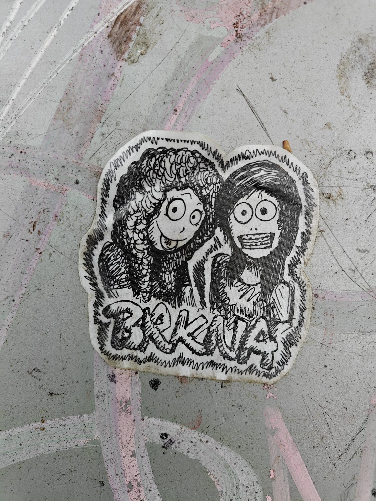 A close-up photograph displays a hand-drawn sticker affixed to a weathered, grey concrete surface with visible layers of faded pink and white paint, and some dirt and scratches. The sticker features two cartoon-like female figures rendered in black and white with a textured, scribbled style.
The figure on the left has large, curly hair, wide, round eyes, and a broad smile with her tongue peeking out, conveying a mischievous or playful expression. The figure on the right has long, straight hair, extremely wide, round eyes, and a very wide, open mouth revealing a full set of teeth, giving her a startled or maniacally cheerful appearance. Both characters are drawn with exaggerated features and a distinct, rough sketch quality.
Below the two figures, the stylized text "BRKNA!" is visible, drawn with a similar hand-scribbled aesthetic. The sticker itself is irregularly cut around the drawing's outline and shows signs of wear, with slight discoloration along its edges, indicating it has been exposed to the elements. The background surface, possibly a wall or urban fixture in Tbilisi, Georgia, is mottled with various textures and faded paint marks, suggesting an aged urban environment. No specific activity is depicted beyond the presence of the street art itself.