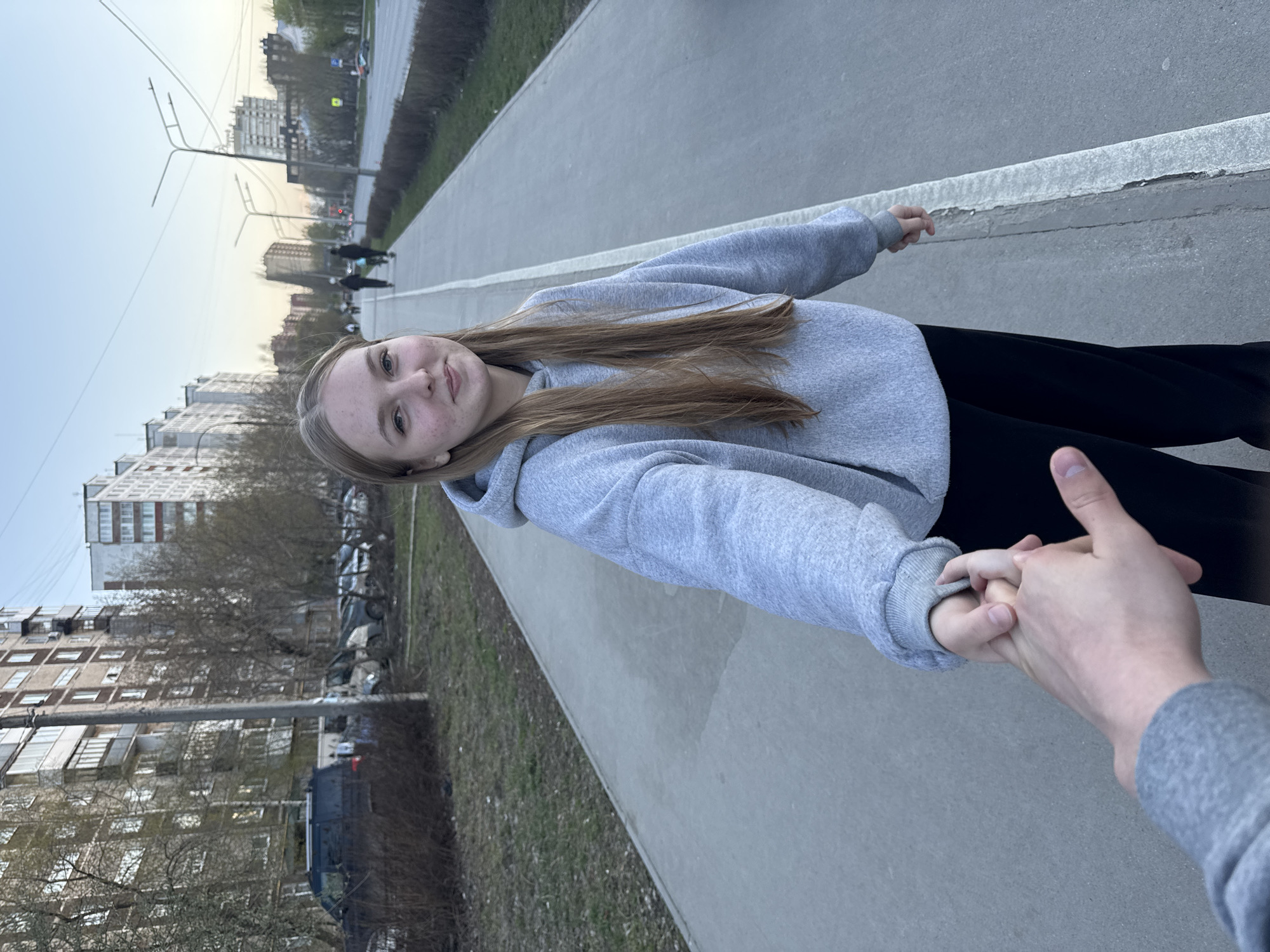 The image depicts a young woman walking alongside an individual, holding hands, in the city of Perm, Russia. The woman is fair-skinned with light brown, shoulder-length hair and freckles. She is wearing a grey hooded sweatshirt and black pants. She is facing the camera with a slight smile. The individual holding her hand is wearing a grey sweatshirt or jacket; only their hand and sleeve are visible.
The setting appears to be a sidewalk or paved walkway adjacent to a street, with buildings, trees, and parked vehicles visible in the background. The buildings suggest an urban residential area. The walkway has a white stripe separating it from the grass. Overhead light fixtures or power lines are visible. The overall scene indicates an overcast day with a late afternoon or early evening light.