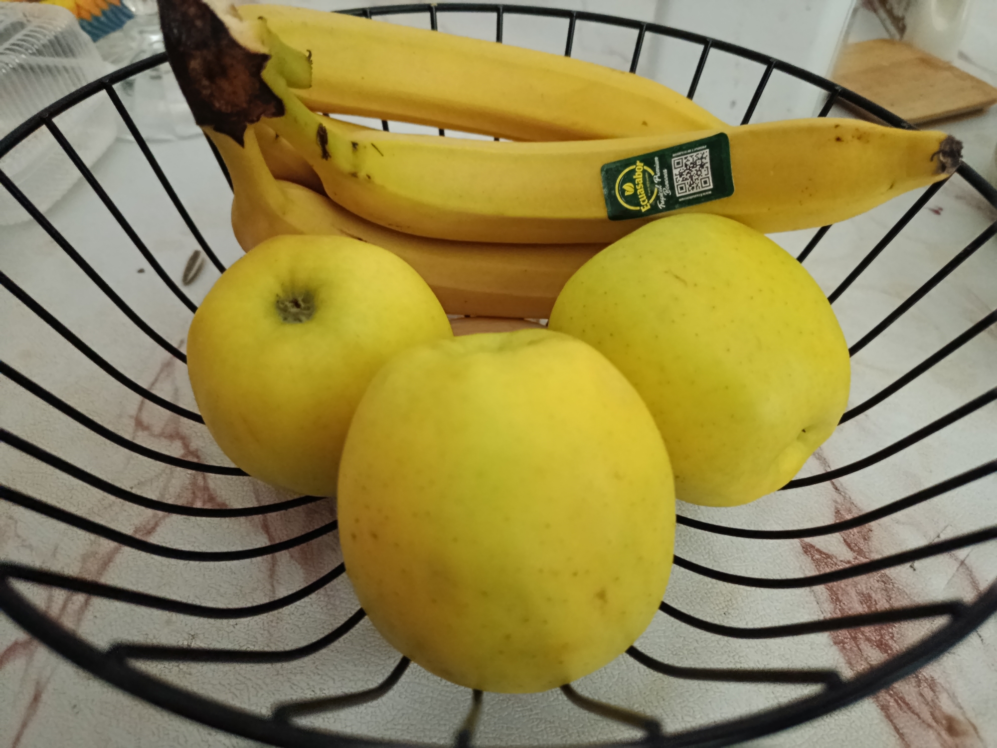 A black wire fruit basket sits on a patterned surface, filled with yellow fruit. Three round, yellow apples are in the foreground, with one in the center and two to its left and right. Behind the apples, a bunch of ripe yellow bananas is visible. The bananas have some brown spots and a green sticker with "Ecuasabor Tropical Premium Bananas" written on it. The surface beneath the basket has a marble-like pattern.