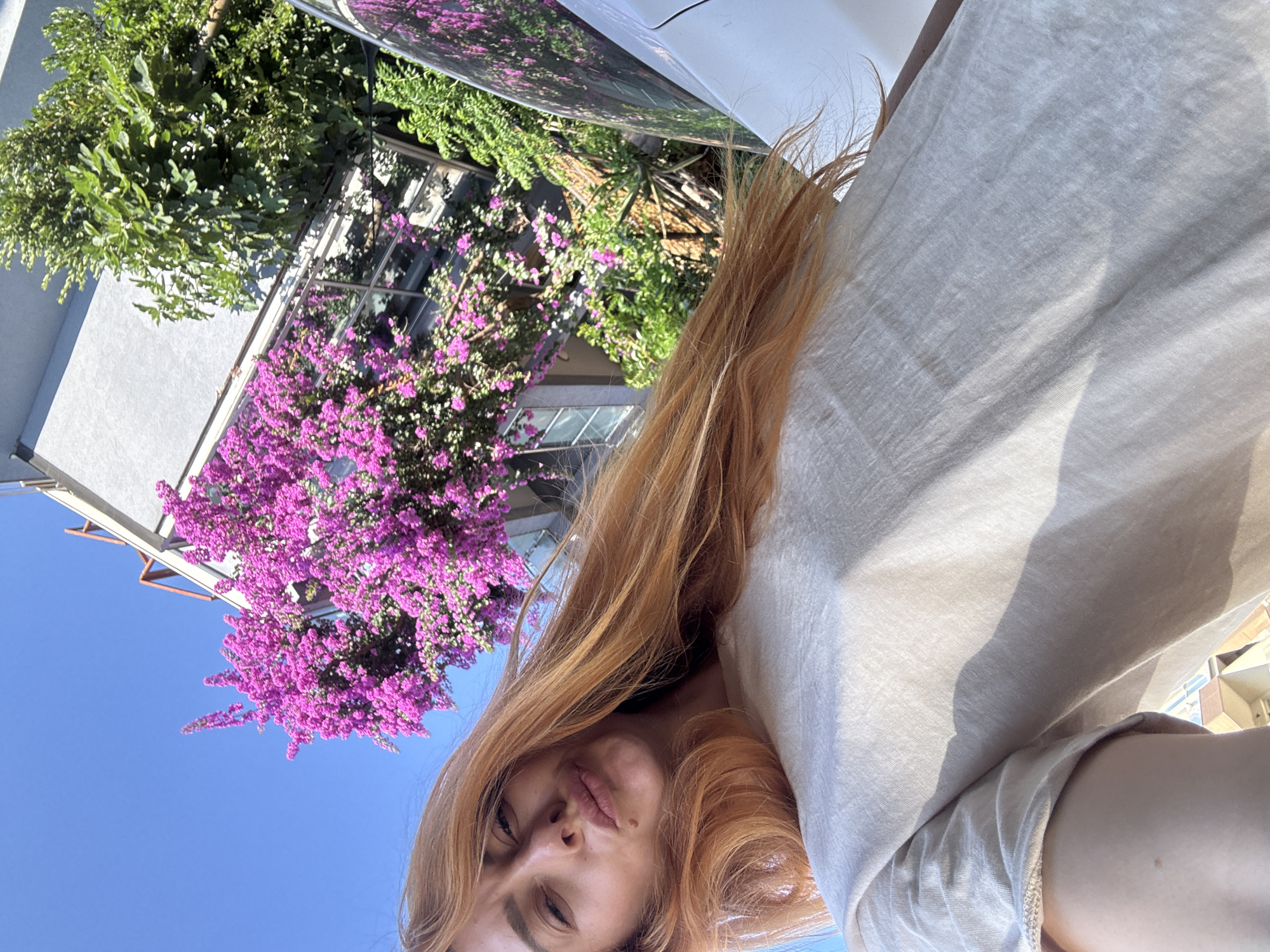 A selfie of a young woman with long, reddish-blonde hair is taken outdoors with a building and vibrant pink bougainvillea flowers in the background. The woman is making a kissing face and wearing a light-colored t-shirt. The sunlight suggests it is daytime. The scene appears to be in Budva, Montenegro, given the characteristic architecture and lush Mediterranean vegetation.