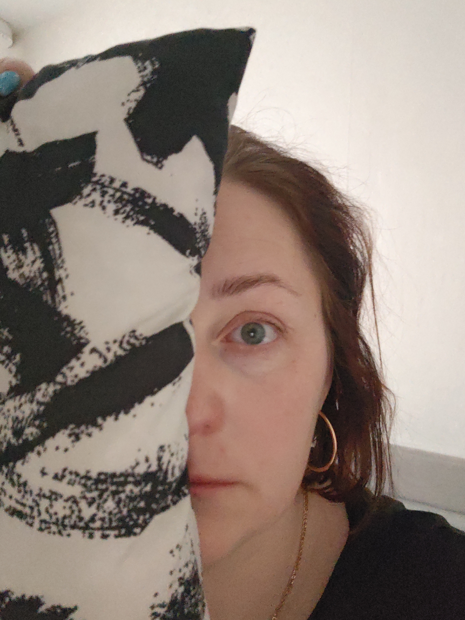 A woman with fair skin and auburn hair is partially obscured by a pillow with a black and white abstract design. Her left eye, which appears green, is visible. She wears a gold hoop earring and a gold chain necklace. She is wearing a black shirt. The background is a plain white wall. The location is not discernible from the image.