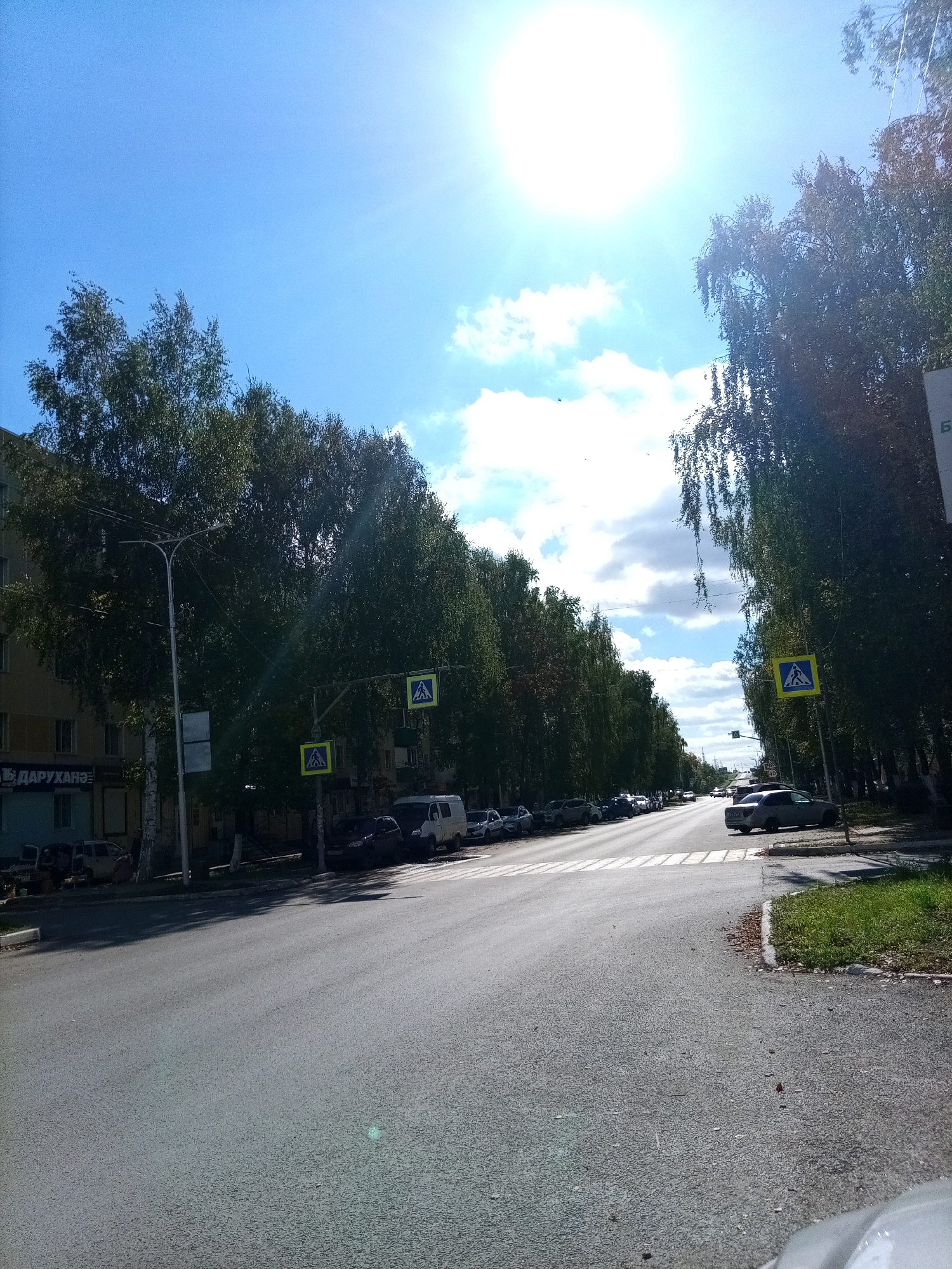 The image shows a sunny day in Aznakayevo, Russia. A street is lined with trees on both sides, with a building visible on the left. There are several cars parked along the street and on the road. Pedestrian crossings are marked with yellow signs. The sky is blue with some clouds, and the sun is shining brightly.