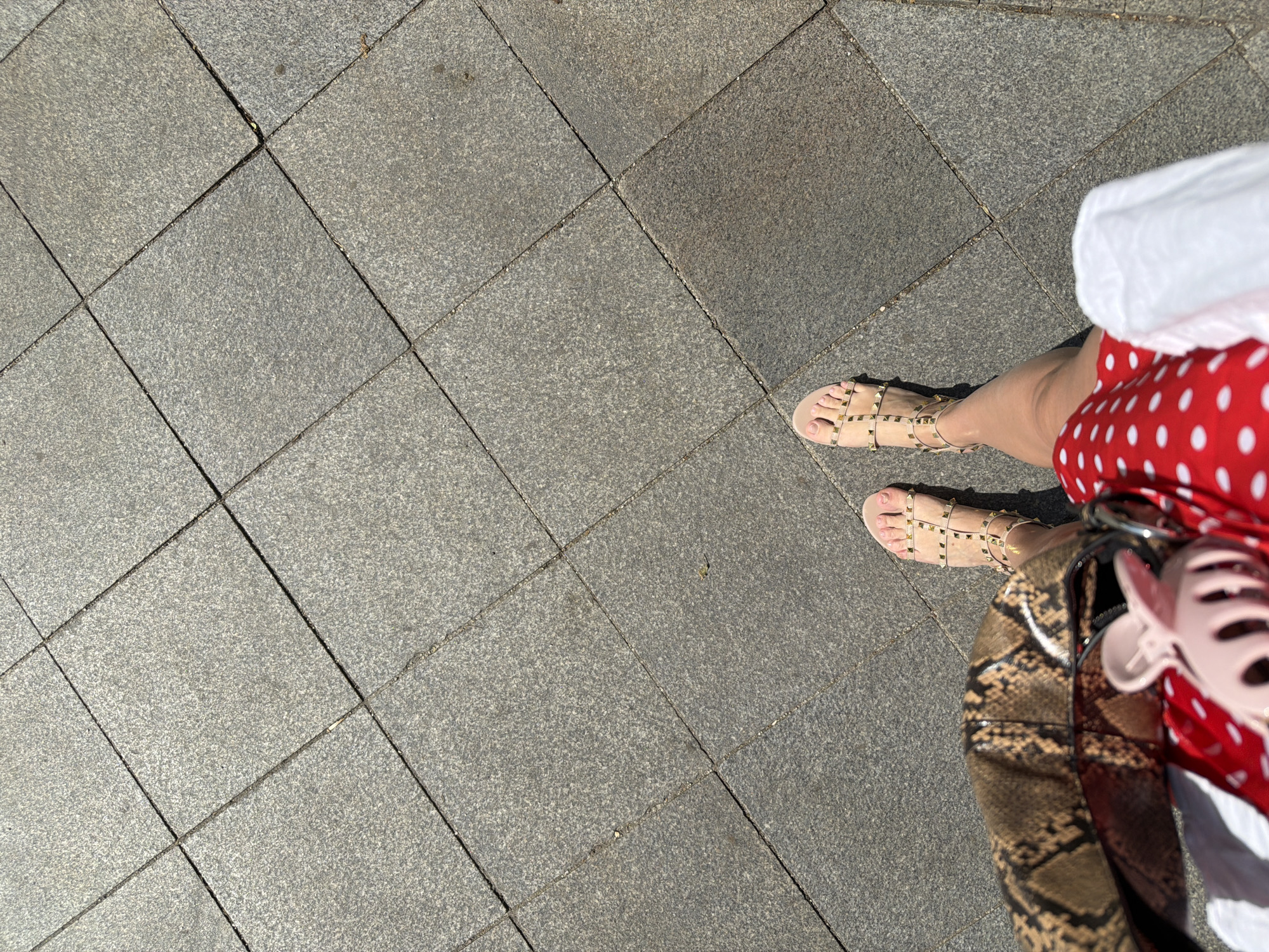 The image presents a first-person, top-down perspective of an individual standing on a paved surface in Moscow, Russia. The pavement consists of square gray tiles. The person is wearing nude-colored sandals with decorative studs, a red and white polka dot skirt, and a white top, with a snakeskin-patterned handbag visible. A pink, Mickey Mouse-shaped object is attached to the handbag. The scene is captured in daylight, with some areas cast in shadow.