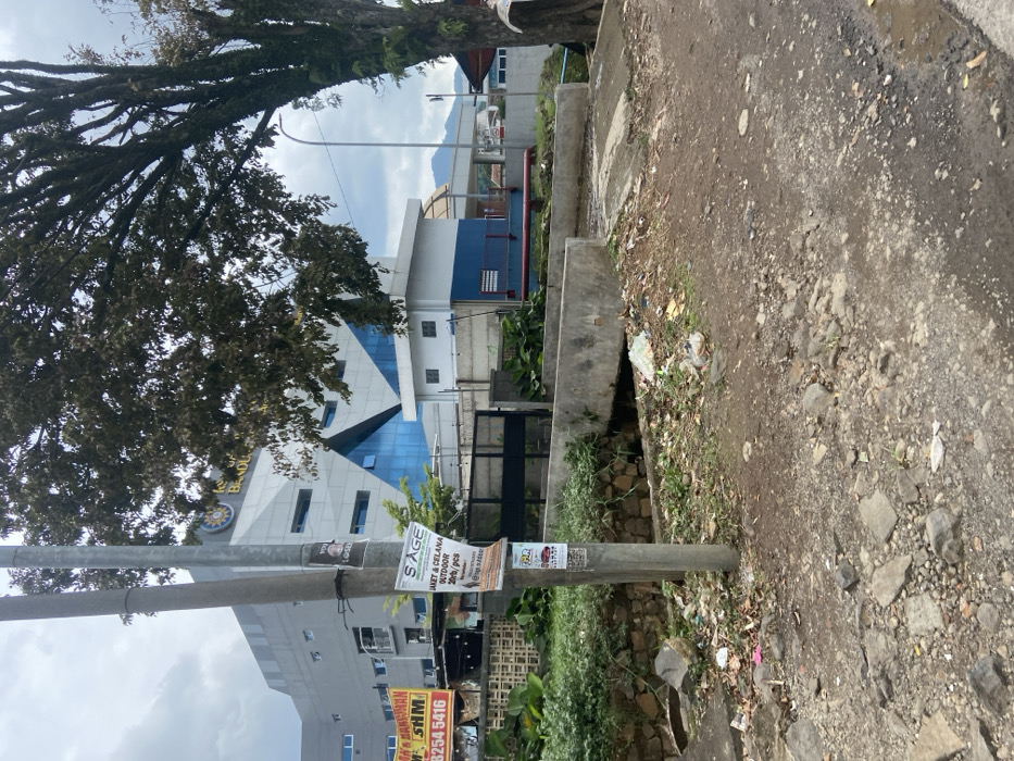 The image shows a street in Ciparay, Indonesia (latitude: -7.022, longitude: 107.68). A concrete bridge spans a small drainage ditch that runs along the street. There is a large tree with dense foliage on the right side of the image. The tree casts a shadow over the bridge and the street. The street is a dirt road with gravel and debris. A gray concrete building with large windows and a blue trim is visible in the background. A large advertisement poster for "SAGE" clothing is attached to a utility pole that supports a street light. The poster is partially obscured by another smaller poster. There is a black metal gate partially visible behind the utility pole. The gate appears to be in the fenced yard of the building. There is an area of dense foliage surrounding the ditch on the left side of the image. The area is littered with garbage and debris. There are no people visible in the scene.