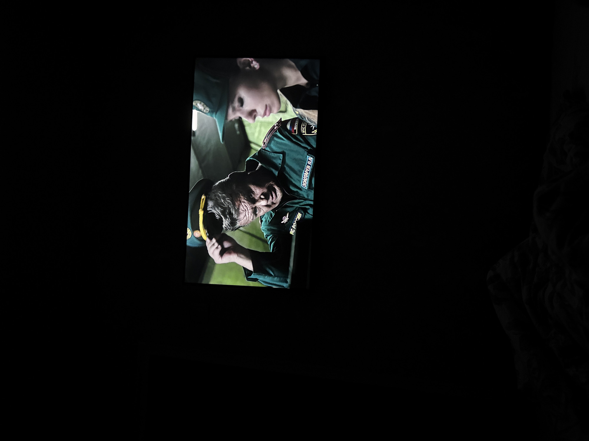 The image displays a screen showing a close-up of two individuals. On the left is a middle-aged man with graying hair, wearing a green uniform with patches and text. He is tipping his hat, revealing a concerned expression. To his right is a young woman, also in uniform, with dark hair and light makeup, looking downwards with a neutral expression. The uniforms appear to be from some sort of emergency service, possibly the Ministry of Emergency Situations of Russia, indicated by the "RUSSIA" and "MЧC POCCИИ" text on their uniforms. The background on the screen is a muted, somewhat blurry green and grey. The overall image is dimly lit, with the screen occupying the central portion and the surrounding area being black. The scene is viewed from a slight angle, and the bottom of the image is predominantly black with some shadowy shapes suggesting a dark room and possibly furniture or a blanket. There is no discernible urban environment or specific location within a city.