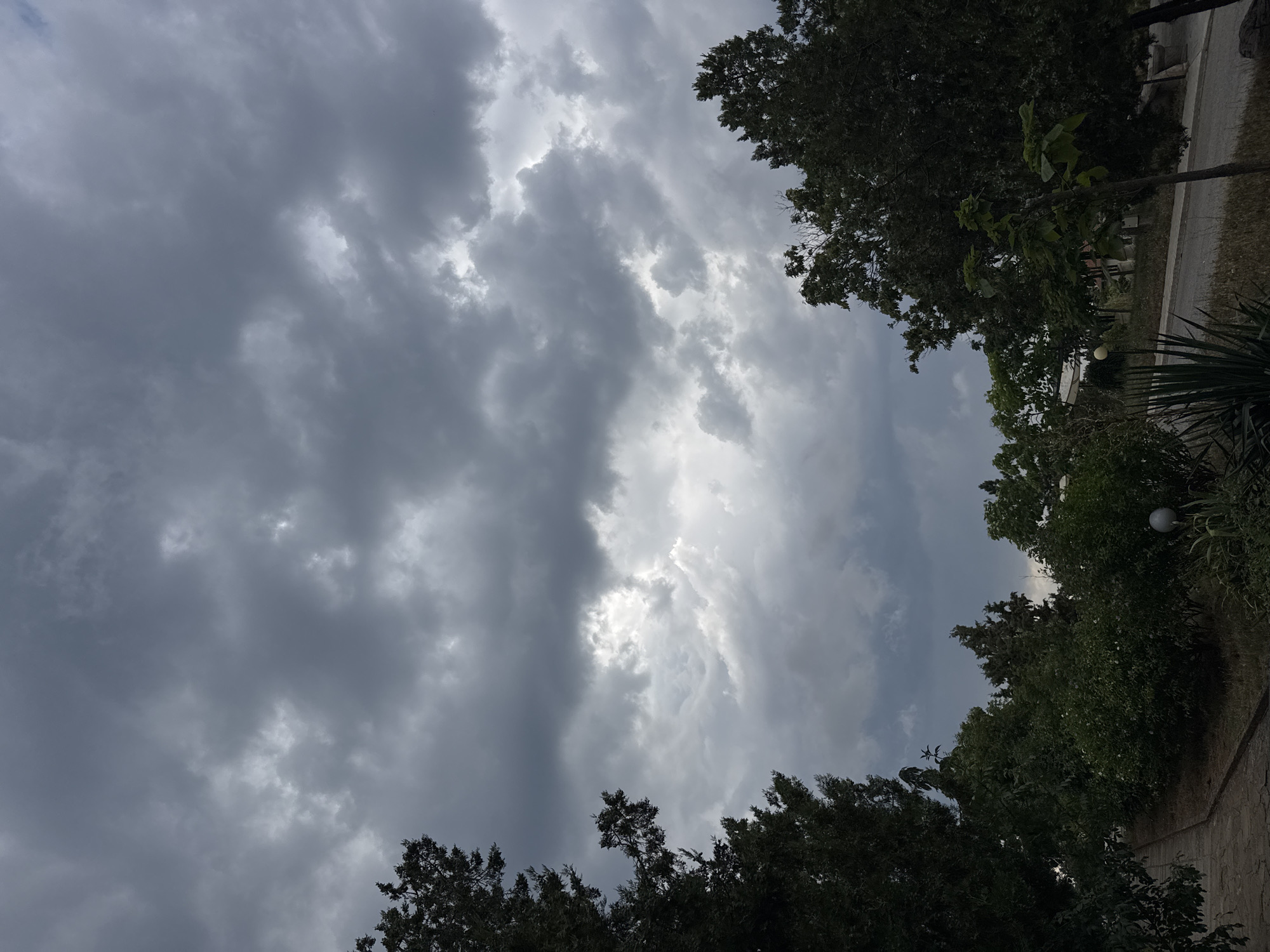 The image shows a cloudy sky over a park-like area in Sredets, Bulgaria. Dark, heavy clouds fill the sky, with patches of lighter clouds suggesting potential breaks in the overcast. Evergreen trees and deciduous trees line a paved walkway in the foreground, leading into a grassy area with some dry vegetation. A street runs along the right side of the park, and a lamppost is visible near the walkway. The overall impression is of a somber, overcast day.