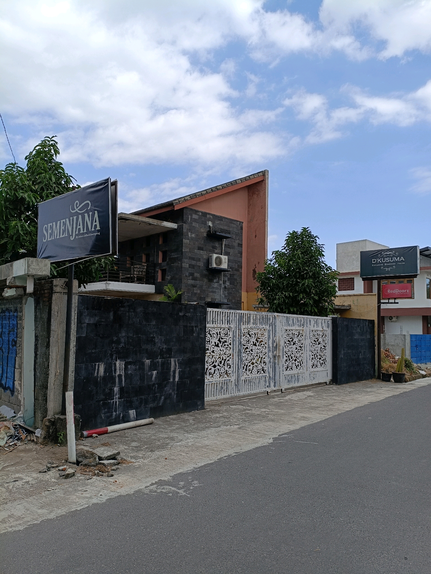 The image shows a street view in Maguwoharjo, Indonesia, featuring a property with a black tiled wall and a decorative white gate. A prominent sign on the left displays "SEMENJANA" in white lettering against a black background, with smaller text below indicating "by Jethro Coffee & Souvenir". To the right, another sign reads "D'KUSUMA EXCLUSIVE BOARDING HOUSE" in white on a black background. Below this is a red sign for "RedDoorZ". The sky is partly cloudy. The road in the foreground is asphalt, and there are some rocks and debris near a concrete pole on the sidewalk.