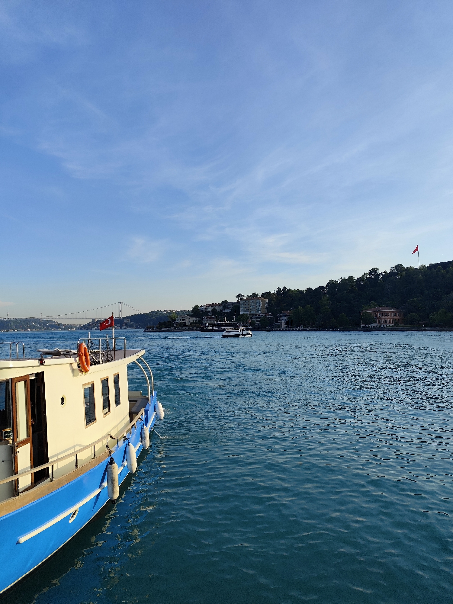 The image presents a serene waterscape viewed from the deck of a boat, likely in the Bosphorus Strait near Sarıyer, Turkey. In the foreground, the starboard side of a white and blue vessel is prominent, featuring a cabin with windows, a wooden railing, and several white fenders hanging along its hull. A small red Turkish flag with a white star and crescent is seen flying from a pole at the boat's stern.
The calm, deep blue water stretches across the mid-ground, showing subtle ripples and reflections. In the distance, a verdant, hilly coastline rises, densely covered with trees and interspersed with various buildings, showcasing a mix of modern and traditional architecture. High on the distant hillside, a large red Turkish flag flies prominently on a flagpole, mirroring the flag on the foreground boat. Further along the horizon, a large suspension bridge, characteristic of Istanbul's Bosphorus bridges, is visible, connecting the distant landmasses. Several other smaller boats and ferries can be seen navigating the waterways in the distance.
The sky above is a clear, bright blue with delicate, wispy white clouds, suggesting fine weather. The quality of light indicates it is daytime, possibly morning or late afternoon, casting gentle illumination on the scene. The overall impression is one of peaceful travel and picturesque scenery, typical of the Bosphorus. No explicit text is visible on any objects beyond the flag emblems.