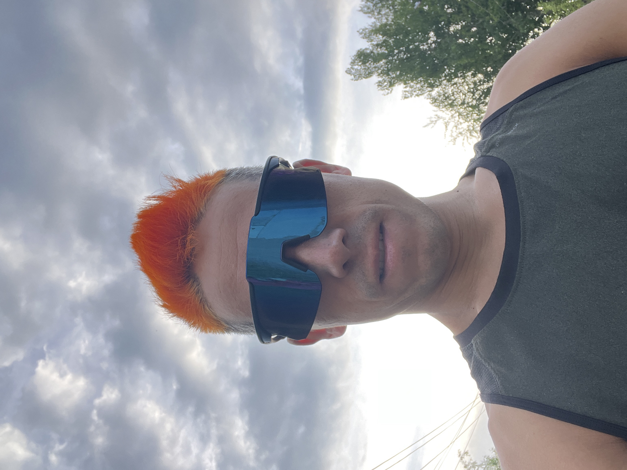 A close-up, low-angle shot depicts a person wearing dark wraparound sunglasses with mirrored lenses. The person has short, orange-dyed hair. They are wearing a dark green tank top with black trim around the neck and arms. The background shows a cloudy sky and the top of a tree. The location is Chaikovsky, Russia.