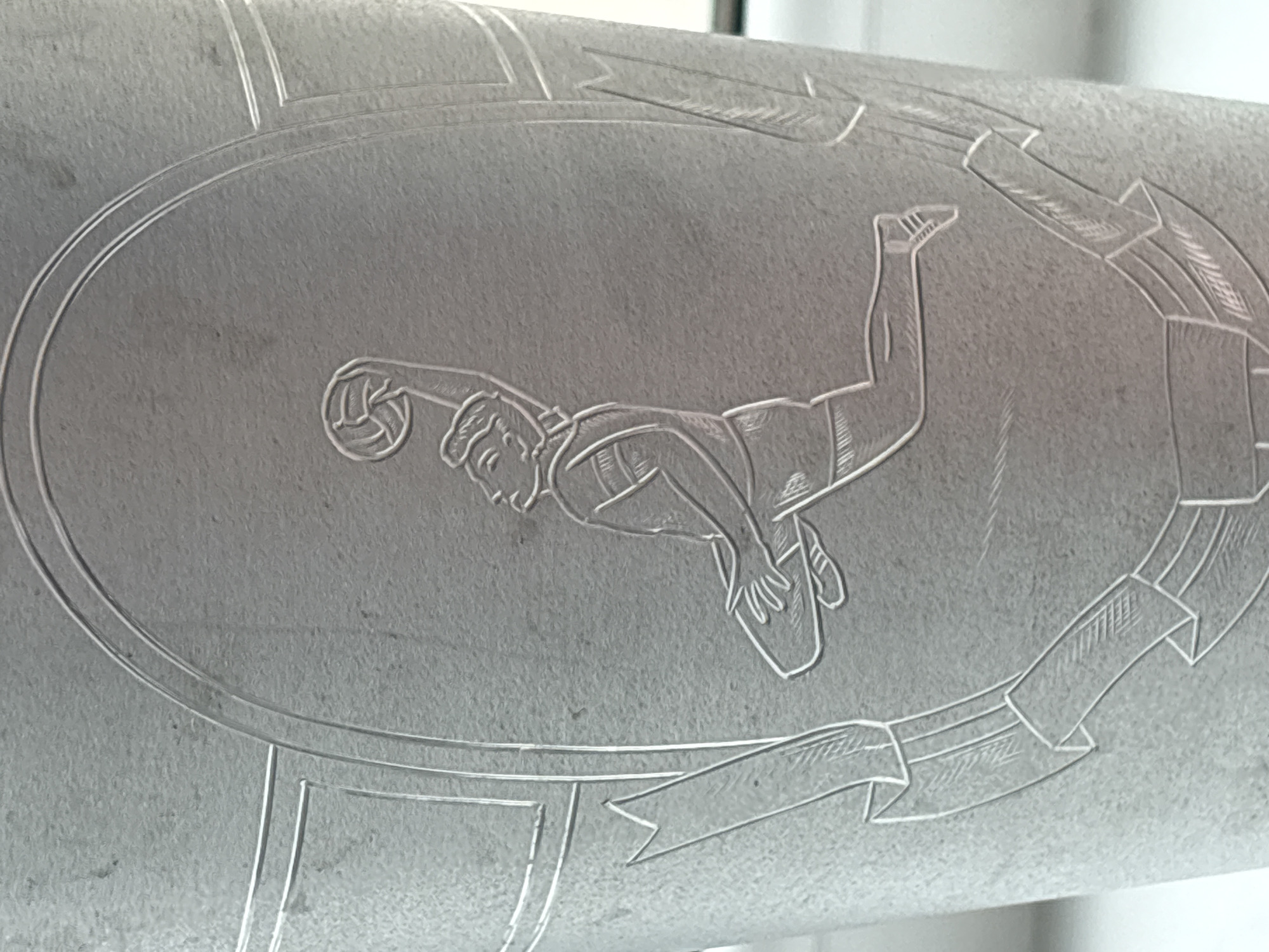 The image shows an engraving, possibly on a metallic surface, depicting a volleyball player mid-dive, reaching for a volleyball with one hand. The figure is surrounded by an oval border and a ribbon-like design. The engraving appears to be relatively detailed, showing the athlete's features, uniform details, and the textured surface of the volleyball. There is no immediate visible indication connecting this image to Izhevsk, Russia.