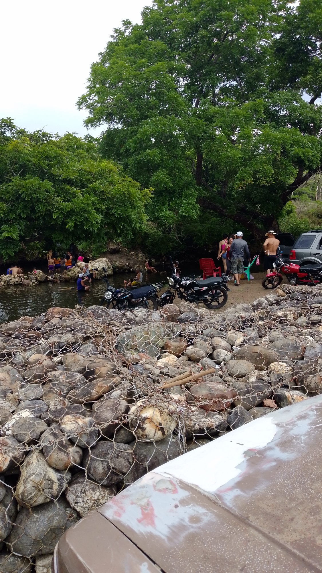 The scene is in Tinaquillo, Venezuela, near a river. A group of people are gathered. Some are in the river, others are on the riverbank. Three motorcycles are parked next to a red plastic chair. A car is parked behind the group. A rock barrier held together by wire mesh lines the riverbank. The sky is overcast, and trees surround the area. The hood of another vehicle is visible in the foreground.