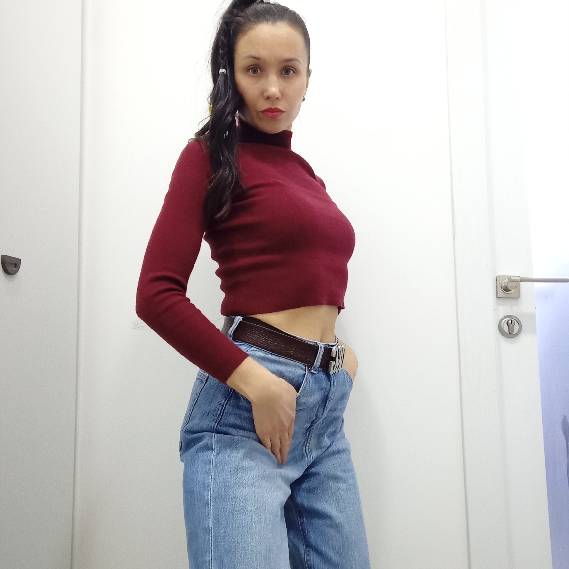 A woman poses indoors against a clean white background, which appears to be a wall formed by two adjacent white doors or panels. She has dark hair styled in a high ponytail with some strands falling forward, and she looks directly at the viewer with a serious or neutral expression, wearing red lipstick. She is dressed in a maroon, long-sleeved, high-neck crop top that reveals her midriff, paired with light blue, high-waisted jeans. A dark brown belt with a prominent silver buckle, featuring what appears to be stylized letters or an unidentifiable monogram, is worn around her waist. Her right hand is casually placed in the front pocket of her jeans.
To the left of the woman, a dark, D-shaped door handle or latch is visible on the white surface. To her right, a modern silver door handle and a keyhole are present on the white door, with a hint of a light blue or purple surface with faint dark markings appearing on the far right edge of the frame, possibly another door or a decorative element. The lighting is bright and even, consistent with an indoor setting. This image was taken in Izhevsk, Russia.