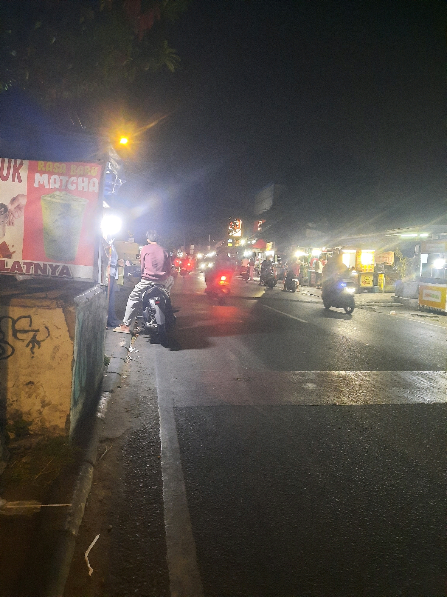 A vertical, medium shot captures a street scene at night in North Jakarta, Indonesia. A person wearing a maroon long-sleeved shirt and grey sweatpants is seated on a black motorcycle, facing away from the camera. The motorcycle is parked on the side of a road with a concrete curb. Adjacent to the motorcycle is a stall with a large poster advertising "RASA BARU MATCHA." To the right of the scene, several other motorcycles are visible, some with riders, and illuminated food stalls line the street. The road surface appears wet, reflecting the lights from the stalls and passing vehicles. The overall atmosphere is that of a busy, nocturnal urban environment.
