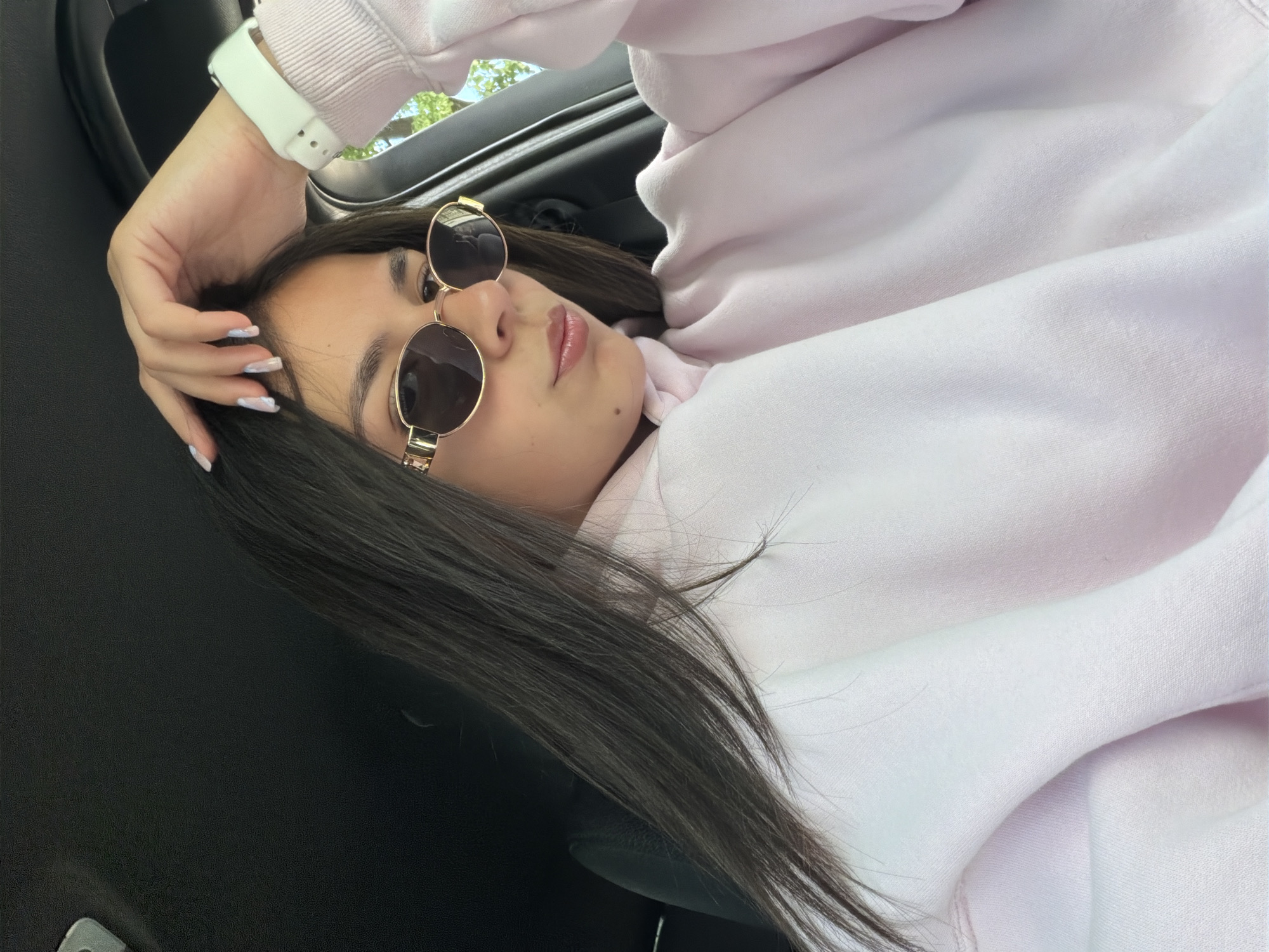 A woman is lying down inside a car. She wears a light pink hoodie and round, gold-framed sunglasses. Her long dark hair is partly spread out on the black leather car seat. She has a white smartwatch on her left wrist. Her hand with manicured nails rests near her forehead. The location is unknown, and there is no information to place it in Gabrovo, Bulgaria.