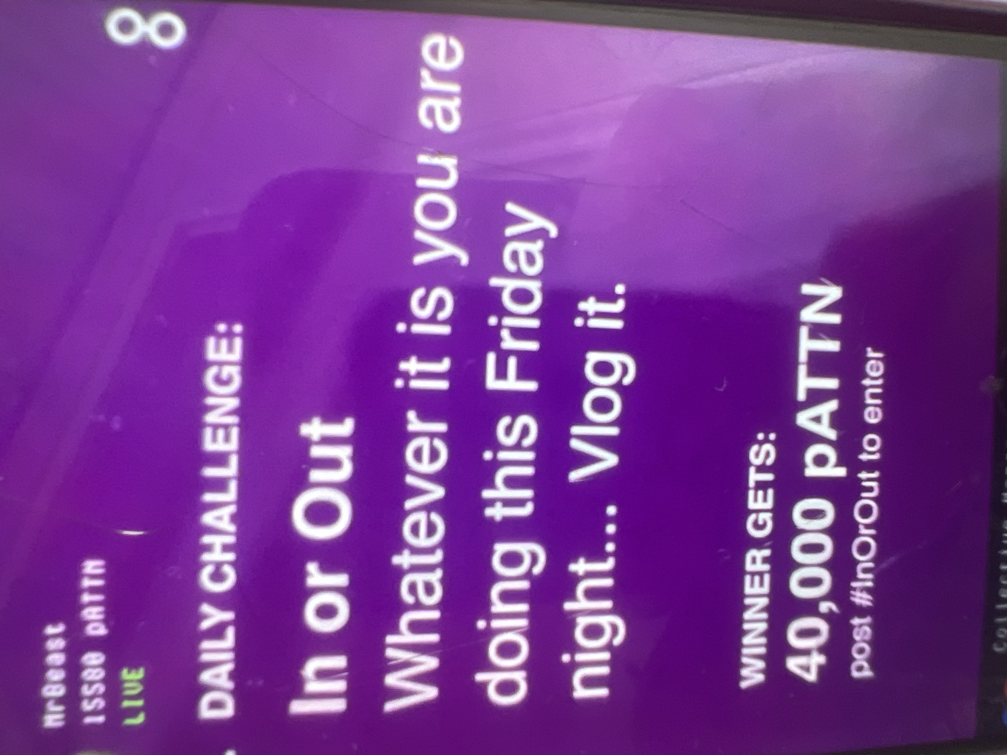 This media file is a photograph of a smartphone screen displaying a live stream or announcement. The dominant color scheme is vibrant purple, suggesting a digital interface or theme.
The screen displays text related to a "DAILY CHALLENGE: In or Out". Below this, a message describes the challenge: "Whatever it is you are doing this Friday night... Vlog it." Further down, it announces that the "WINNER GETS: 40,000 PATTN" and instructs viewers to "post #InOrOut to enter".
In the bottom left corner, there is information about the streamer or channel, reading "MrBeast", "15580 PATTH", and "LIVE" in green text. An infinity symbol "8" is visible in the top right corner of the screen.
The photo has a slightly distorted perspective, indicating it was taken from an angle of a phone screen, possibly held by a person. The lighting is bright, and there are some visible reflections and light streaks on the screen, suggesting the photo was taken in a well-lit environment, possibly indoors. The purple background is consistent across the screen, with the text in white. The overall impression is of an interactive online event or promotion.