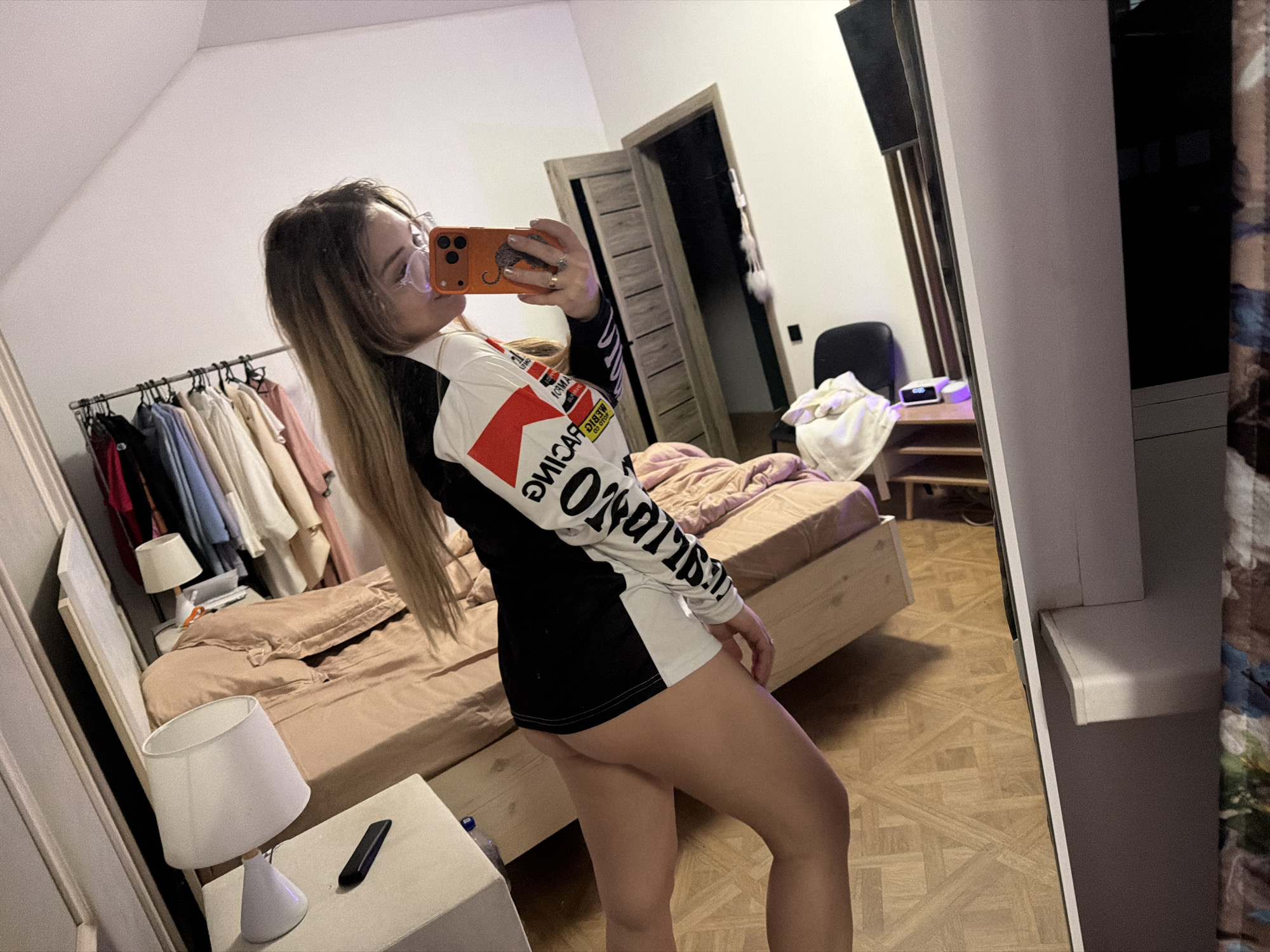 A young woman is captured in a mirror selfie within a bedroom setting. She stands with her back partially turned to the camera, holding an orange smartphone to take the photo. Her long, straight blonde hair with darker roots falls over her shoulders, and she wears clear-rimmed glasses. She is dressed in an oversized, long-sleeved top that is black on the body and white on the sleeves, featuring racing-themed graphics. Visible text on her shirt, reversed due to the mirror, includes what appears to be the "MARLBORO" logo in a red block, and "RACING" along with other sponsor names like "MOTUL," "PIRELLI," and "CHAMPION" vertically arranged on the white sleeve. Her bare legs are visible from mid-thigh down.
The bedroom features light-colored walls and a parquet-style wooden floor. To the left, a metal clothing rack holds various garments. A wooden bed frame with light brown bedding occupies the center-left. Two white nightstands flank the bed; one closer to the viewer has a white lamp and a black remote control, while another further back also holds a lamp. In the background, a light brown wooden door is ajar, and a black office chair is visible. A tall, light-colored cabinet or shelf unit stands on the far right, partially obscured by the mirror. A small digital alarm clock is visible on a shelf near the black chair. The overall impression is a casual, domestic scene, likely in Большая Шильна, Russia.