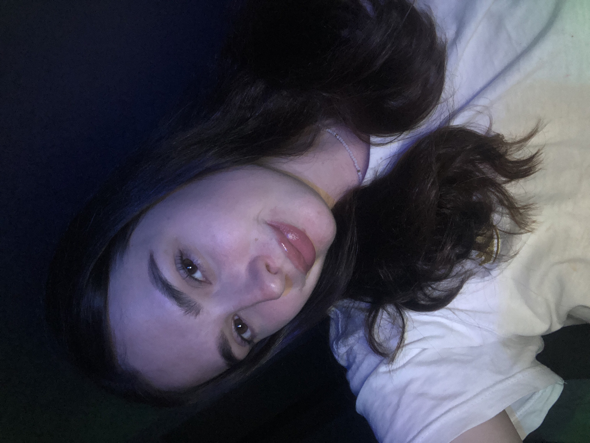 The image shows a close-up, selfie-style shot of a young woman with long, dark brown hair. She is wearing a white t-shirt and a thin, beaded necklace. Her eyes are brown, and she is smiling slightly. The lighting is dim and appears to be from artificial sources, casting a purplish hue on her skin. The background is dark and indistinct, suggesting an indoor setting at night. There is no visible indication of the city of Noviy, Russia in the image.
