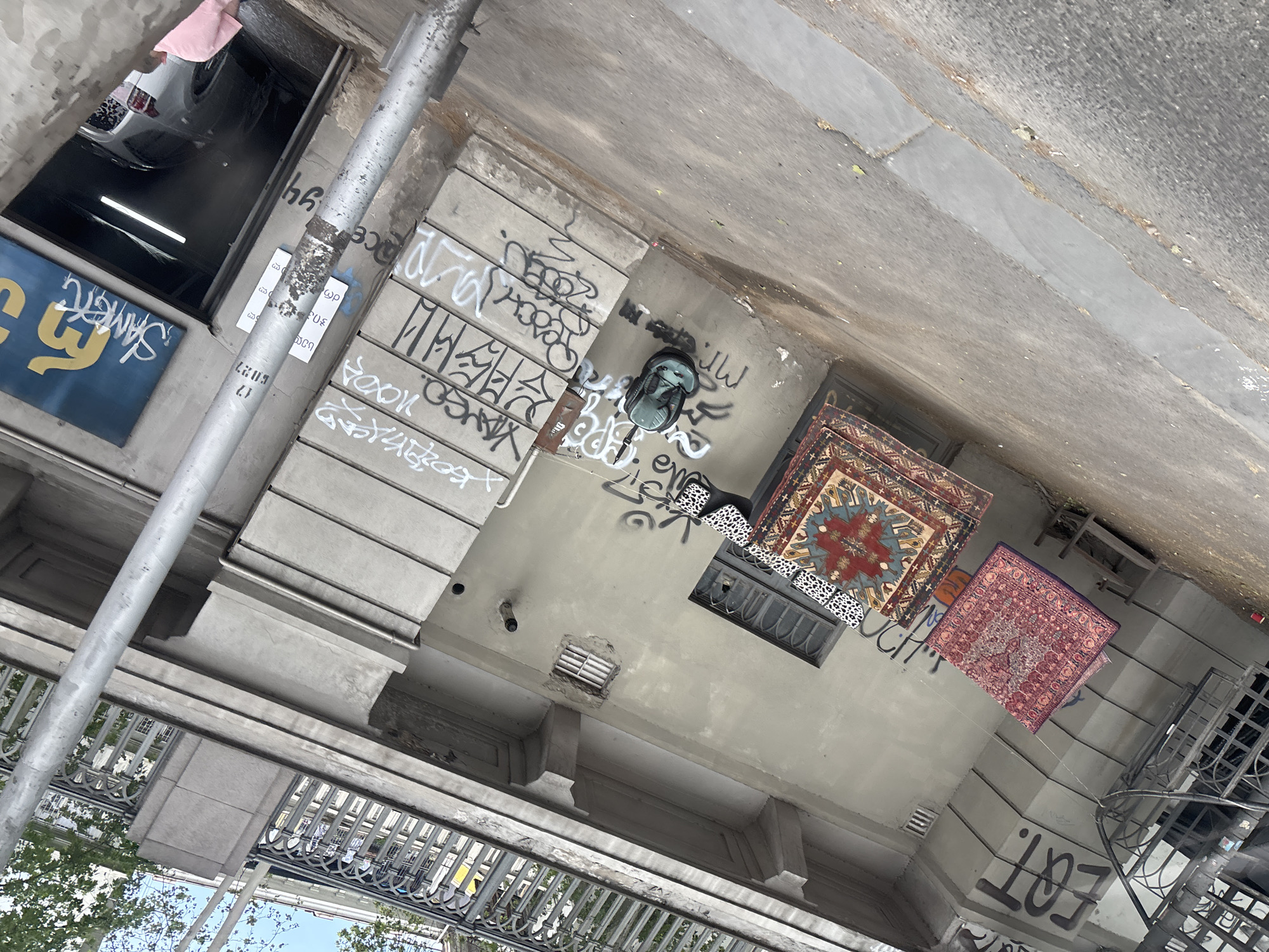 An overhead shot depicts a building exterior in Tbilisi, Georgia, featuring two Persian-style rugs hanging from wires strung between the windows and architectural elements of the building. Graffiti covers much of the exterior surface, including the building’s wall and adjacent column. One of the rugs is a square, primarily red with blue and gold patterns. The other, slightly smaller, appears to be a maroon color. A backpack rests on the surface beneath the rugs, surrounded by additional graffiti. Part of the building's exterior features what looks like black iron bars of a small window. There are tree tops and blue sky visible in the lower left of the frame. A parked car can be seen in the upper left corner.