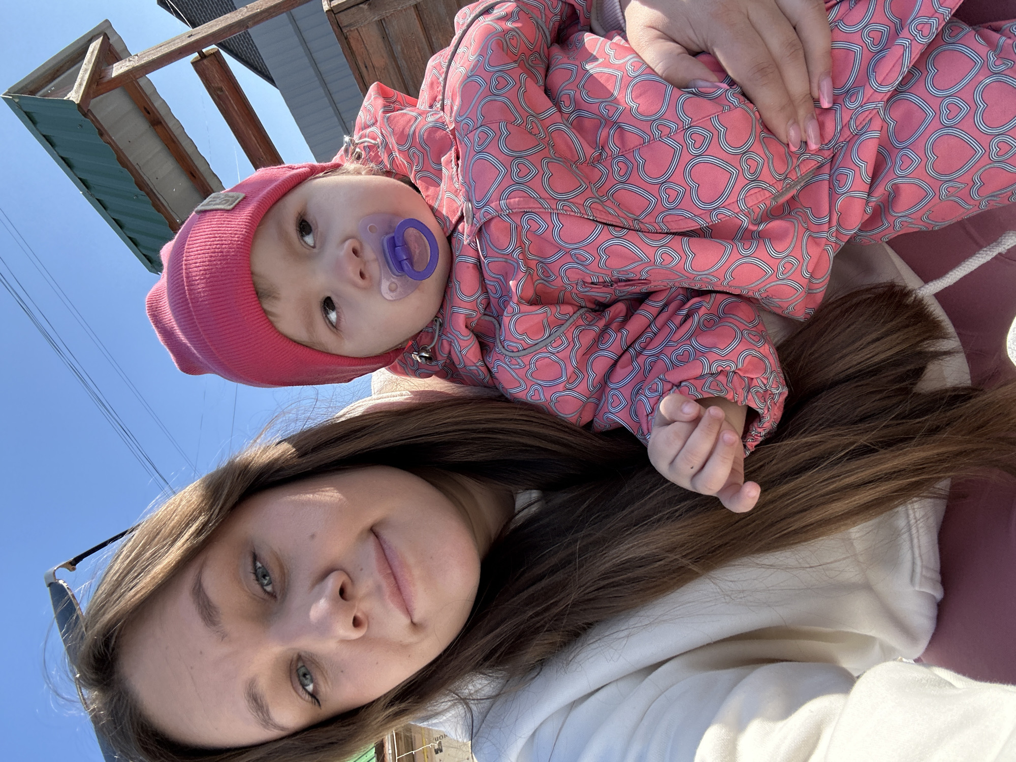 A selfie photograph shows a woman and a baby outdoors. The woman has long brown hair and is wearing a white hoodie. The baby, wearing a pink heart-patterned jacket and a pink hat, has a purple pacifier in its mouth. The baby is being held by the woman. A partial view of a building structure is visible in the background, along with a clear blue sky. The image was taken in Ольховка, Russia.