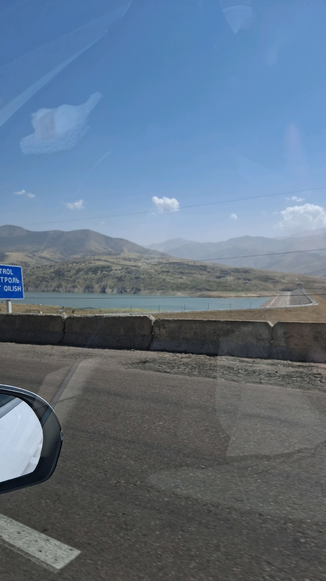 The image, captured from a vehicle, overlooks a body of water nestled within a valley of rolling hills under a clear blue sky with scattered white clouds. In the foreground, the asphalt road and a concrete barrier are visible. To the left, a side-view mirror of the vehicle is reflected. A blue sign with white text, partially obscured and illegible, stands near the road. The landscape appears arid and mountainous, with sparse vegetation on the hillsides. The overall scene suggests a scenic drive through a natural landscape, possibly in a border region given the presence of a control sign. The location is identified as Chinar, Uzbekistan.