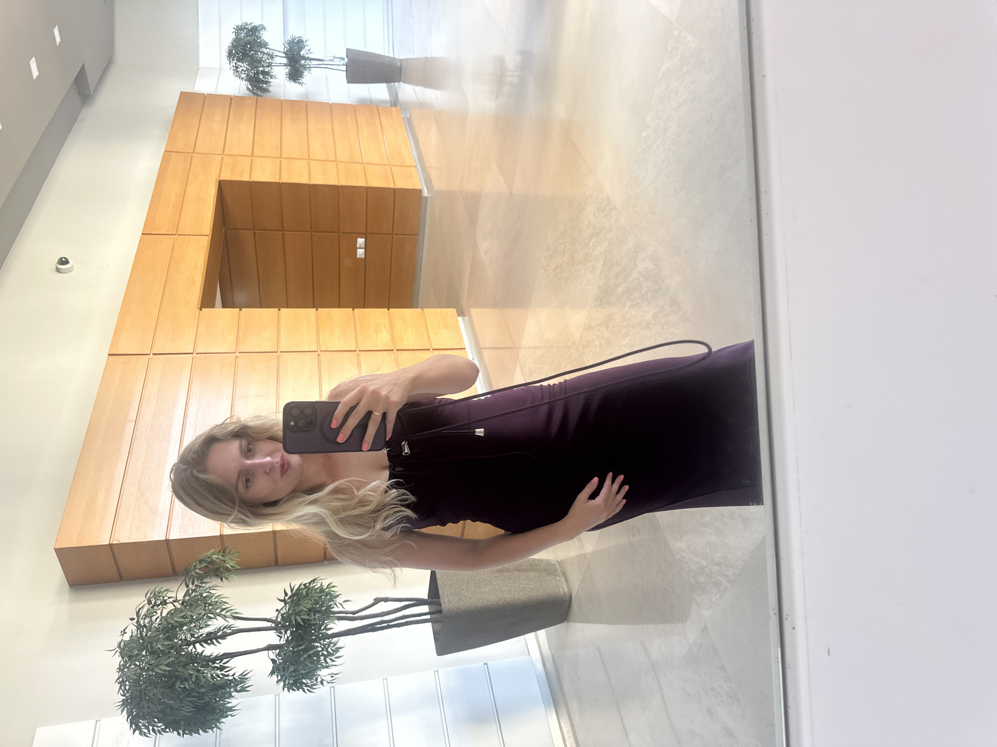 A woman with long blonde hair, wearing a dark purple dress, is taking a mirror selfie in a modern, well-lit interior space. She is holding a dark-colored smartphone in her right hand, with her left hand resting on her hip. The background features a wall clad in light brown wood panels, with a dark opening in the center. To the left and right of the frame, there are tall, narrow planters with artificial green plants. The floor is polished marble or tile, reflecting the light and the objects in the room. The ceiling appears to be white, with recessed square lighting fixtures and a visible security camera. The environment suggests a hotel lobby or a similar upscale public space. This scene is located in the city of Petah Tikva, Israel.