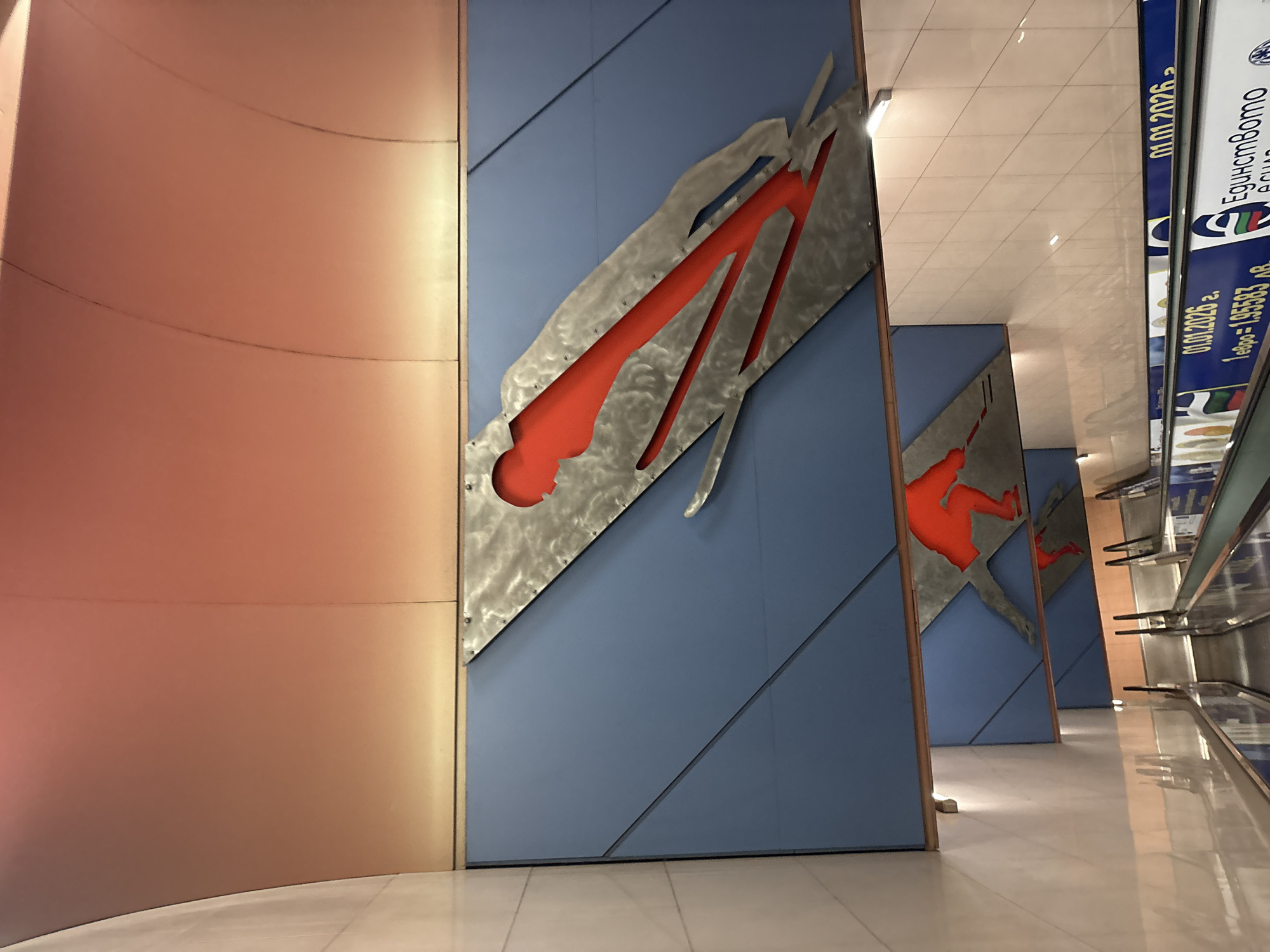 The image depicts an interior space, likely a subway station based on the presence of escalators on the right. Dominating the left and center of the frame are several large wall panels decorated with stylized figures of skiers, indicated by the shape of their bodies and equipment. The panels consist of blue backgrounds with cutout sections filled with red and bordered by metallic silver framing. The first panel on the left is curved and has a brown gradient. The background suggests a modern or recently renovated space, with tiled flooring and bright lighting. The location is likely in Sofia, Bulgaria. The signs on the right are in Bulgarian.