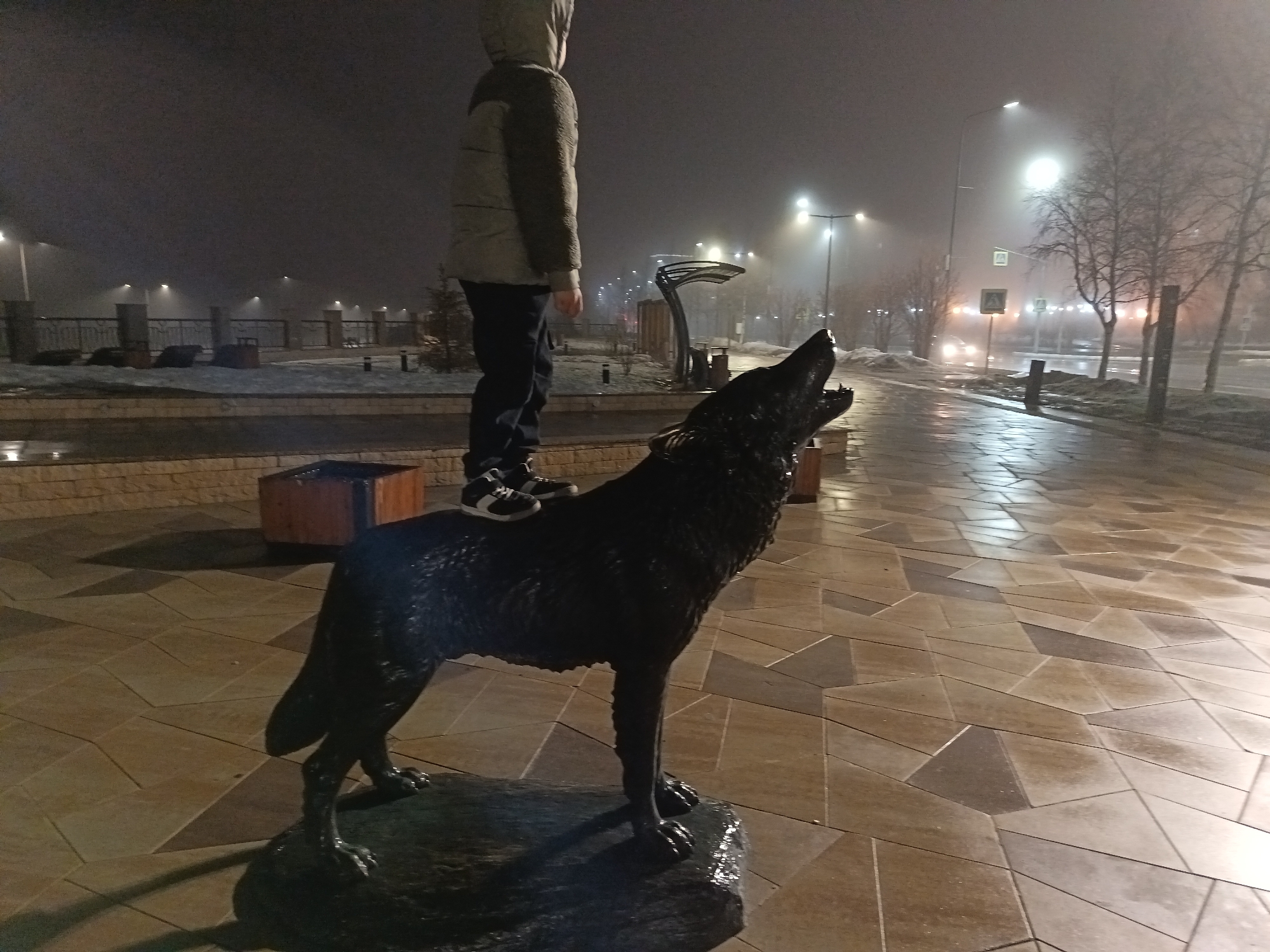 A child stands on a statue of a wolf howling at night in Nizhnevartovsk, Russia. The child is wearing a hooded jacket and dark pants. The statue is black and wet, likely from the damp night. The ground is paved with polygonal stone tiles. In the background, street lights illuminate a foggy street with sparse trees. There's a metal arch sculpture, and other street furniture is visible. Snow is present on the grassy areas. A car's headlights pierce through the fog.