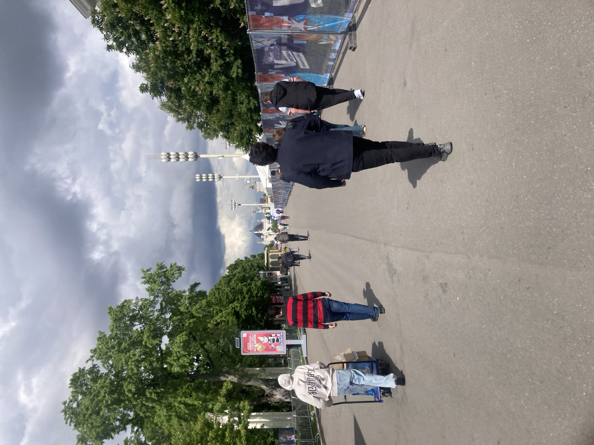 The image depicts a paved walkway in what appears to be a park or pedestrian area. A person in a dark jacket and pants is walking along the walkway. Further down the path are other pedestrians. There are trees on either side of the walkway. The sky is cloudy. Signs with Cyrillic text are visible, suggesting the location is in Russia, likely Moscow, given the presence of distinctive street lights that are known to be present in the city. A person is pulling a trolley with packages. Another person is wearing a red and black striped shirt.