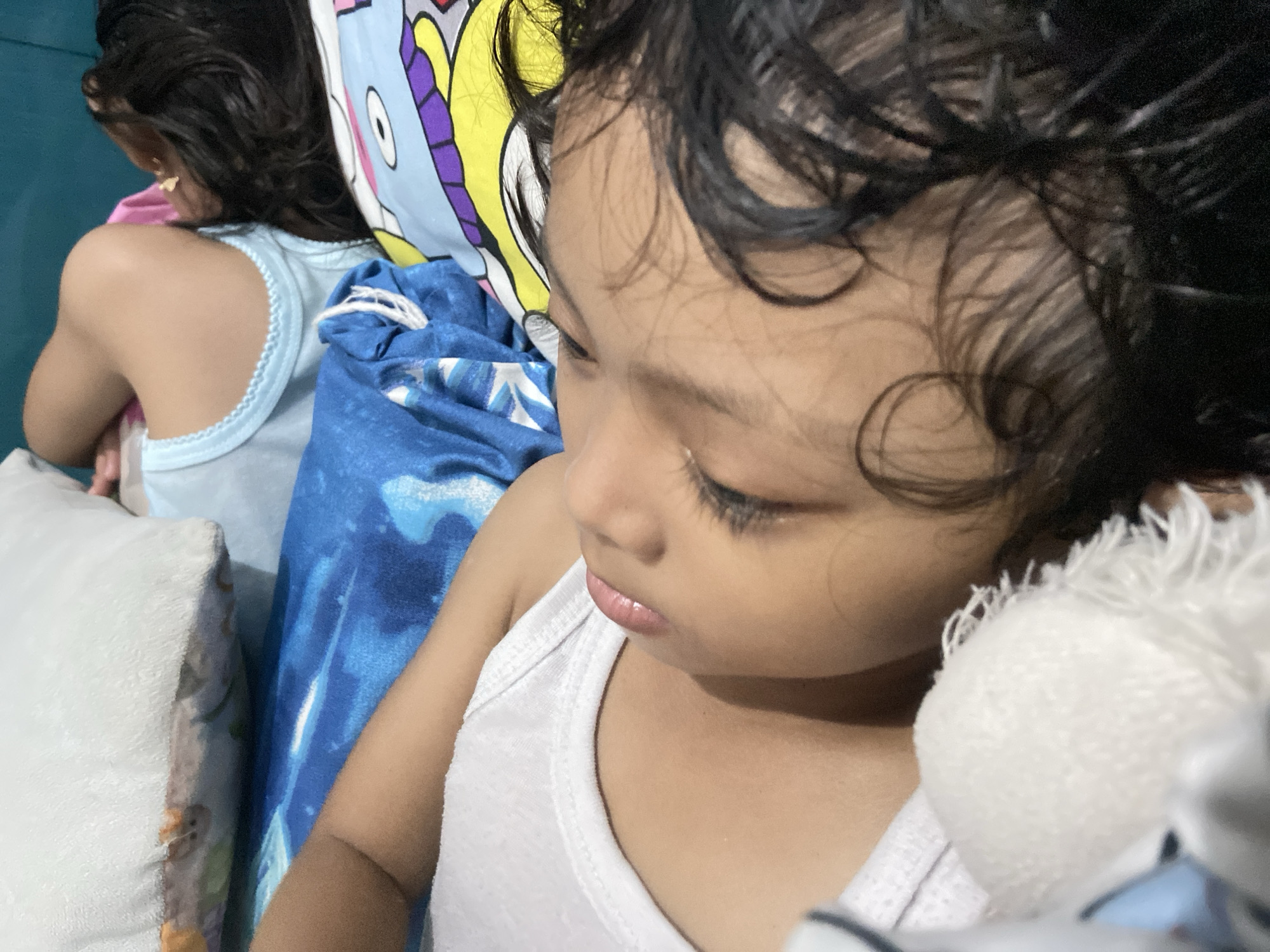 A close-up, indoor shot shows a child lying down with their eyes closed, appearing to be asleep. The child has dark hair and is wearing a white tank top. They are resting on a bed or sleeping surface, with a blue and white patterned blanket visible beneath them. Another person, possibly another child, is partially visible in the background, lying on a pillow with a colorful cartoon design. The pillow is adjacent to the child in the foreground. A stuffed animal is placed near the face of the child in the foreground. The location, Gelam, Indonesia, cannot be confirmed from visual data only.