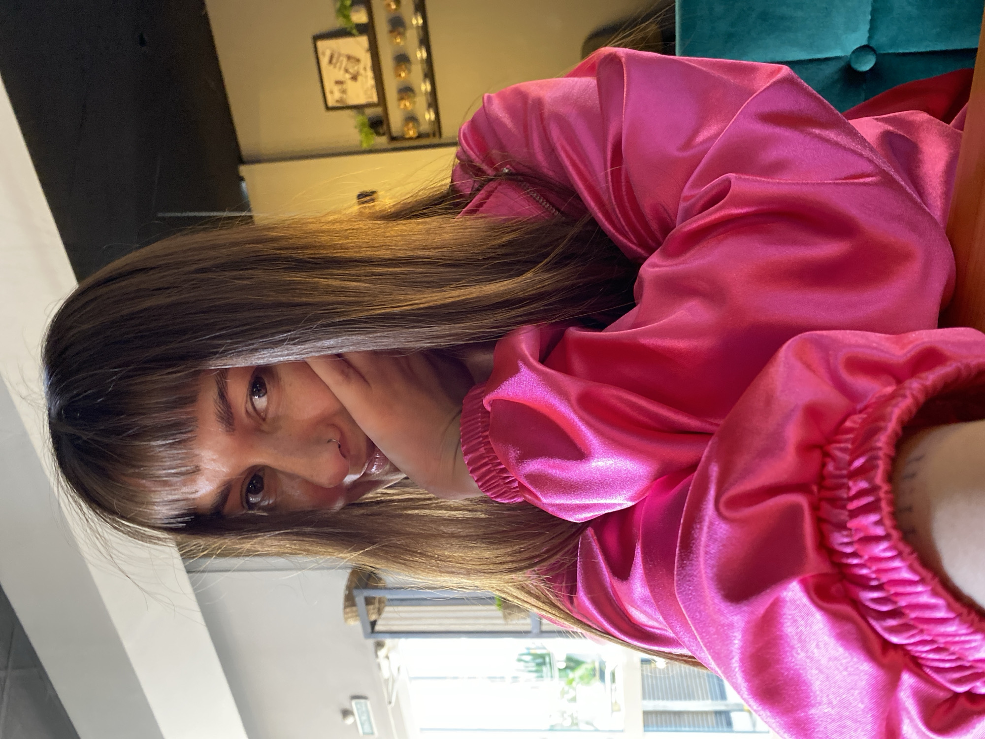 A woman with brown hair and bangs is shown in a close-up, eye-level shot. She is wearing a shiny pink top with elastic around the wrist. Her face is partially obscured by her hand. The background includes a wall with framed artwork and decorative items, a teal cushioned surface, and a window showing an exterior scene. The image is taken at an angle, giving a slightly skewed perspective. There is no indication in the image that this was taken in Votkinsk, Russia.