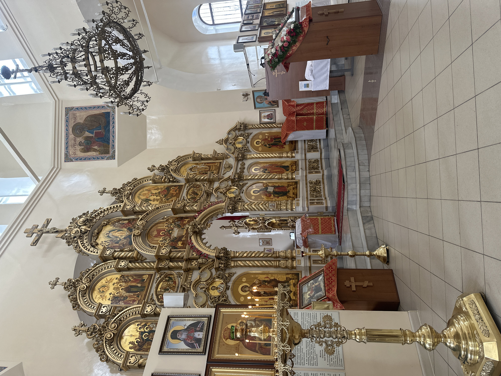 The image shows the interior of what appears to be an Orthodox church. The focal point is a highly ornate iconostasis adorned with numerous icons rendered in gold and vibrant colors. There are several steps leading up to the iconostasis.
In the background, there is a wooden altar or table decorated with flowers and a crucifix. Above the iconostasis, a window provides natural light. A large, ornate chandelier hangs from the ceiling.
To the left of the image, several framed icons can be seen. There are tall golden candle stands in the foreground. The floor is covered with tiles. The location is likely a church in Votkinsk, Russia.