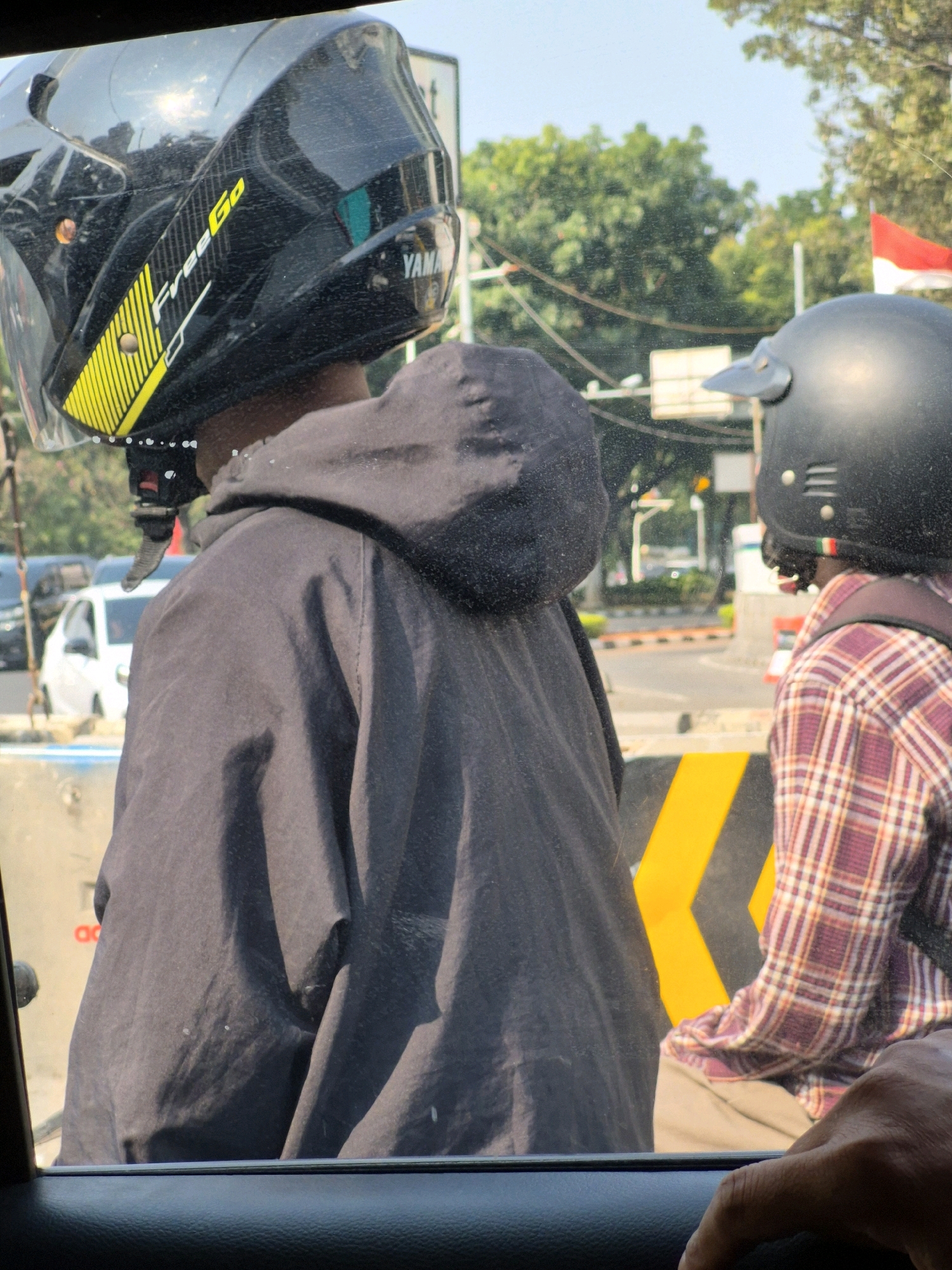 The image captures a scene from inside a vehicle looking out at two individuals on a motorcycle in what appears to be traffic. The person in the foreground is wearing a black helmet with yellow and white decals and the brand name "FreeGo" is visible. They are also wearing a dark colored hooded jacket. Behind them, another individual is wearing a black helmet with a red, white, and green stripe on the side. This person is wearing a red and white plaid shirt and a dark backpack. The background shows trees, traffic signals, a white car, and the Indonesian flag. The environment suggests a sunny day in a city, possibly Central Jakarta, Indonesia, given the presence of traffic and urban elements.