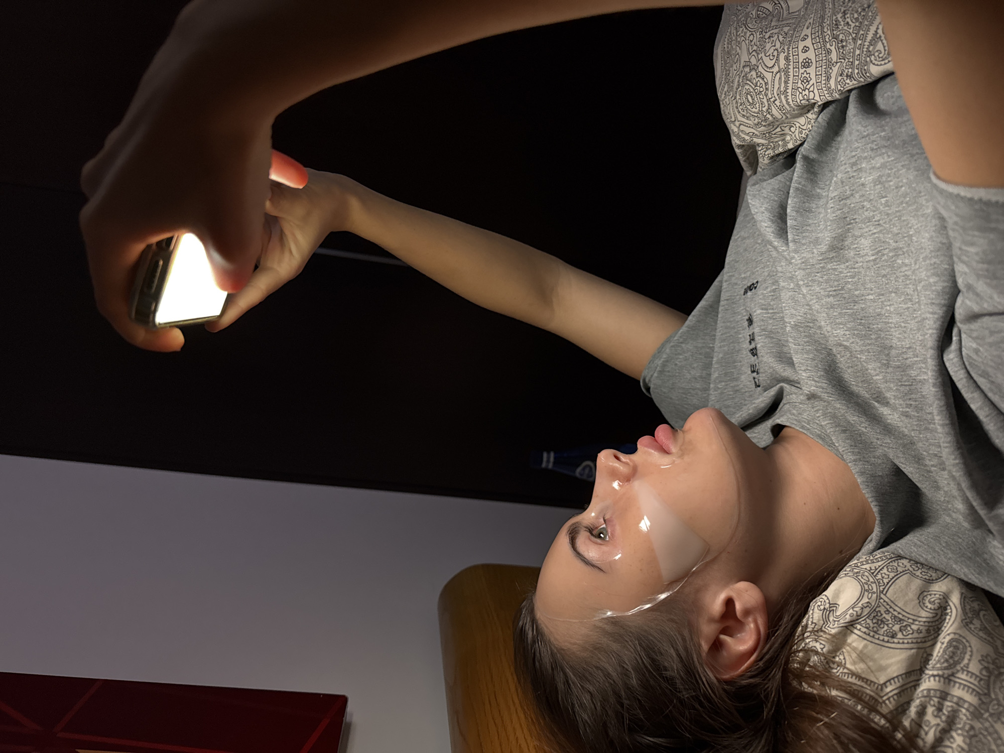 A person lies supine on a patterned pillow, likely in a domestic interior, holding a mobile phone aloft. The phone screen is illuminated, and the person's face, adorned with what appear to be cosmetic strips, is angled towards the light. The environment features a two-tone wall with a dark upper portion and a lighter lower portion. A small section of what may be furniture is visible on the left side of the image. The overall scene suggests a nighttime or low-light setting. There's no evidence to place the image specifically in Warsaw, Poland.