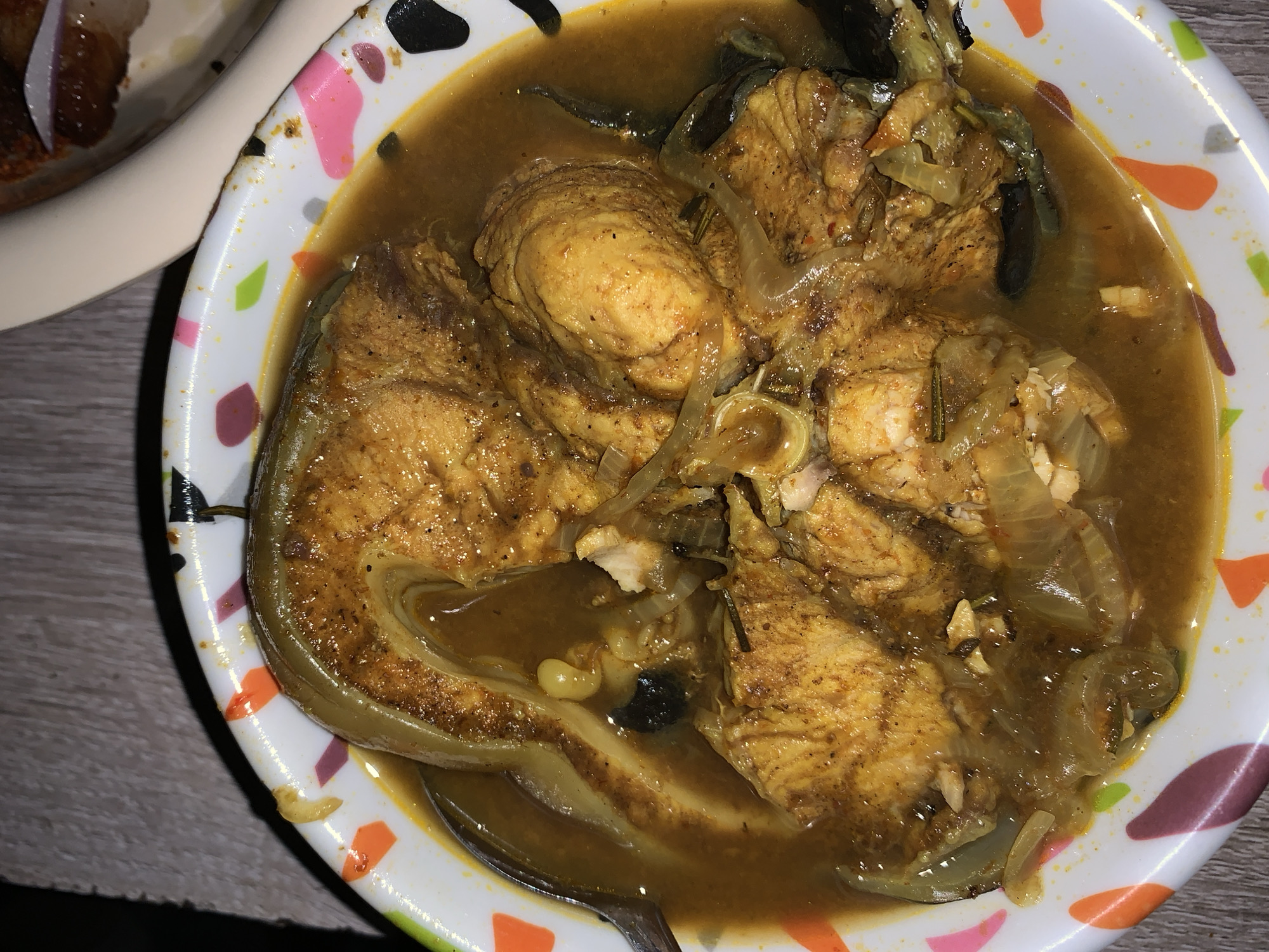 A close-up shot of a plate of stewed fish. The fish is arranged in the center of the plate and surrounded by a brown sauce. The fish is topped with onions. The plate has a patterned rim with shapes. A spoon rests in the sauce. Part of another plate is visible in the top left corner. The background is a dark surface. There is no information about the location or the city of Kabong, Nigeria in the image.