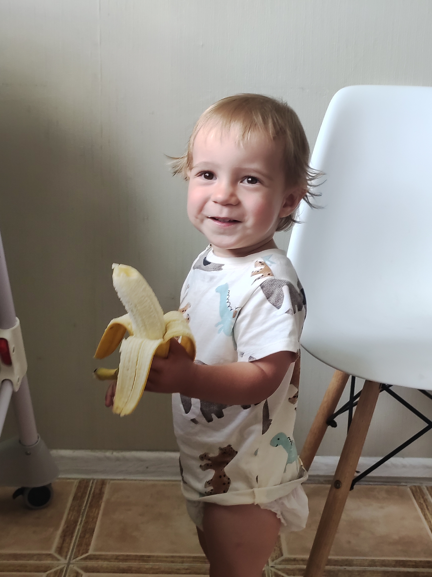 A young child, likely a toddler, stands in front of a plain wall and a white chair, holding a partially peeled banana. The child has light brown hair and is smiling. They are wearing a white t-shirt with a pattern of brown and blue dinosaurs, and a white diaper. The banana is held in the child's right hand, with the peel partially pulled back, revealing the fruit. To the left of the child, part of a gray baby walker with wheels is visible. The floor is tiled in a pattern of beige and brown squares. The lighting suggests it is daytime. The setting appears to be indoors, possibly a home.