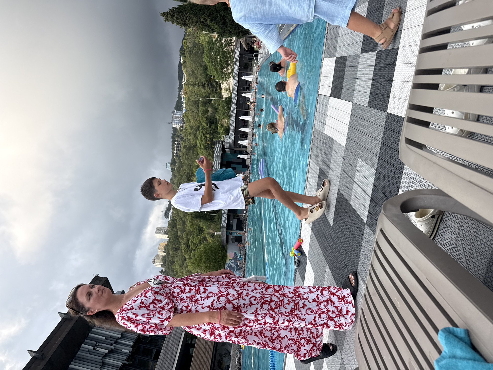 A woman in a red and white floral dress walks on a tiled poolside next to a swimming pool filled with people. A boy in a white t-shirt and patterned shorts walks beside her, holding a purple object. The pool area is surrounded by trees and buildings on a hillside. The sky is overcast. In the foreground, a lounge chair is partially visible. Several people are swimming and floating in the pool. The location is the Yalta city pool.
