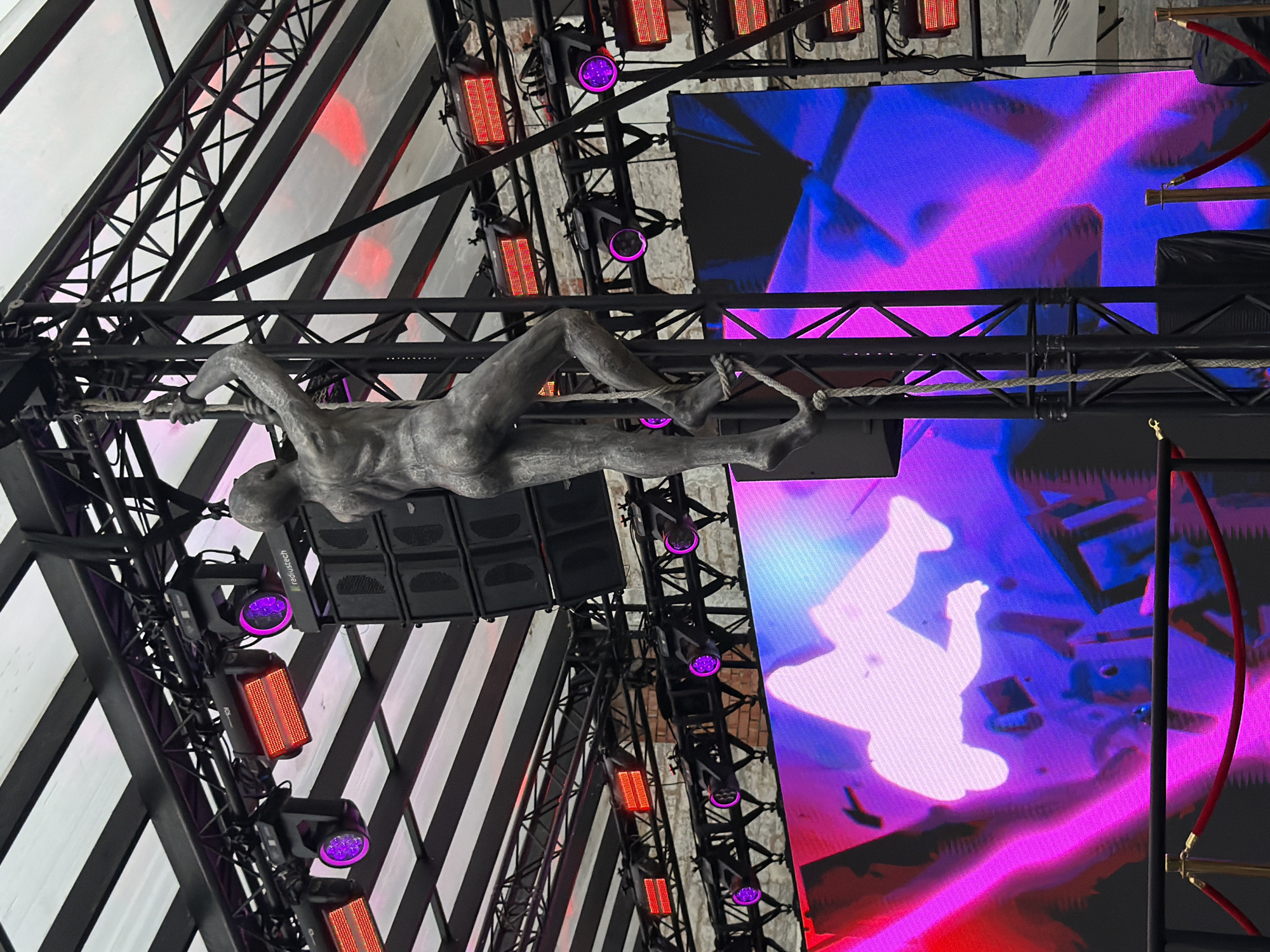 A statue of a man climbing a rope is positioned on a metal structure beneath a white tented roof. The structure is equipped with multiple speakers and stage lights, some emitting purple and red hues. In the background, a large LED screen displays a vibrant pink and blue abstract image, with a silhouette of a person jumping. This scene is located at the Tallinn Song Festival Grounds in Tallinn, Estonia.