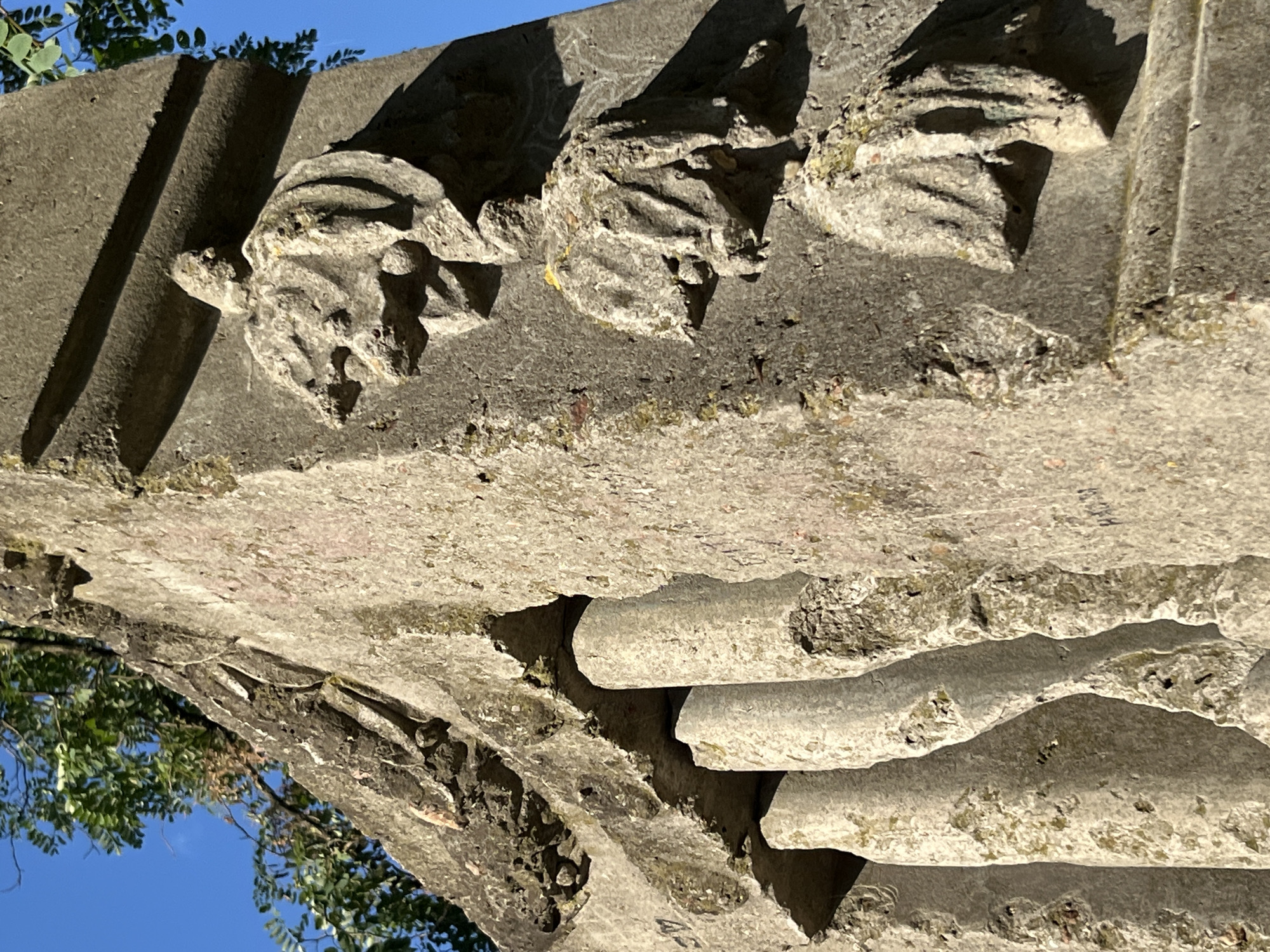 The image shows a close-up view of a weathered stone monument in Narowlya, Belarus. The monument, likely a tombstone or part of a larger structure, features carved reliefs of what appear to be stylized leaves or flower motifs. The stone is rough and pitted, with visible signs of age and erosion. Some green moss or lichen is present on the surface, particularly in crevices. The sun is casting shadows, highlighting the three-dimensional nature of the carvings. In the background, parts of a tree with green leaves and a clear blue sky are visible. The angle of the photograph suggests the monument is oriented towards the sky and is outdoors. The details of the carvings are somewhat obscured by the weathering and the angle, but the general forms are discernible.