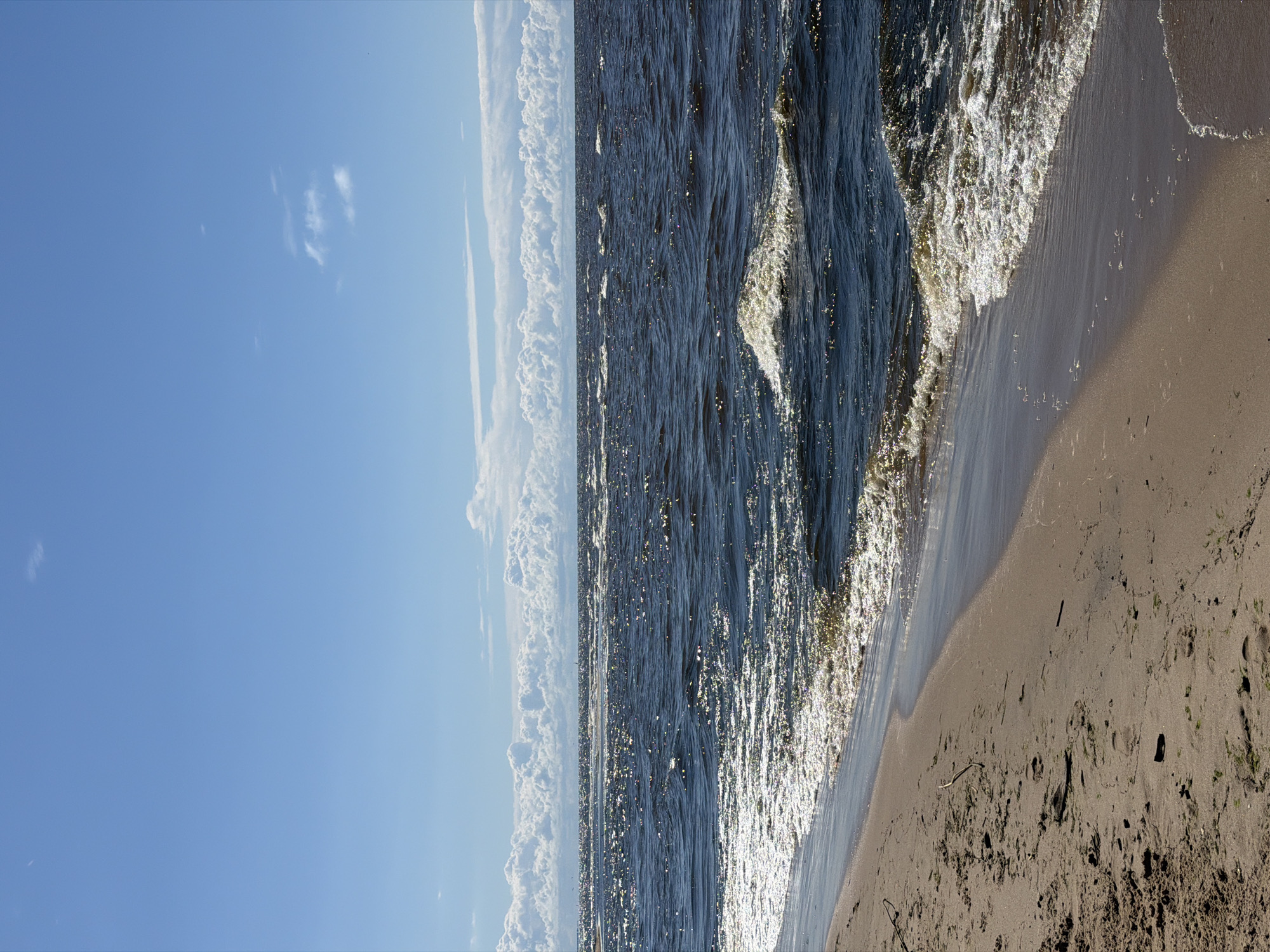 This is a vertical, eye-level, medium shot of a sandy beach meeting a dark, choppy sea under a blue sky with scattered white clouds. The sand is light brown with some small pieces of debris, including twigs and seaweed, and is marked with footprints. The sea is a deep blue, with white foam and cresting waves visible along the shore. Sunlight glints off the surface of the water, creating a shimmering effect. In the distance, along the horizon, there are more white clouds forming a band across the sky. The image captures a natural coastal environment. The location is the beach in Gauja, Latvia. There are no people visible in the image.